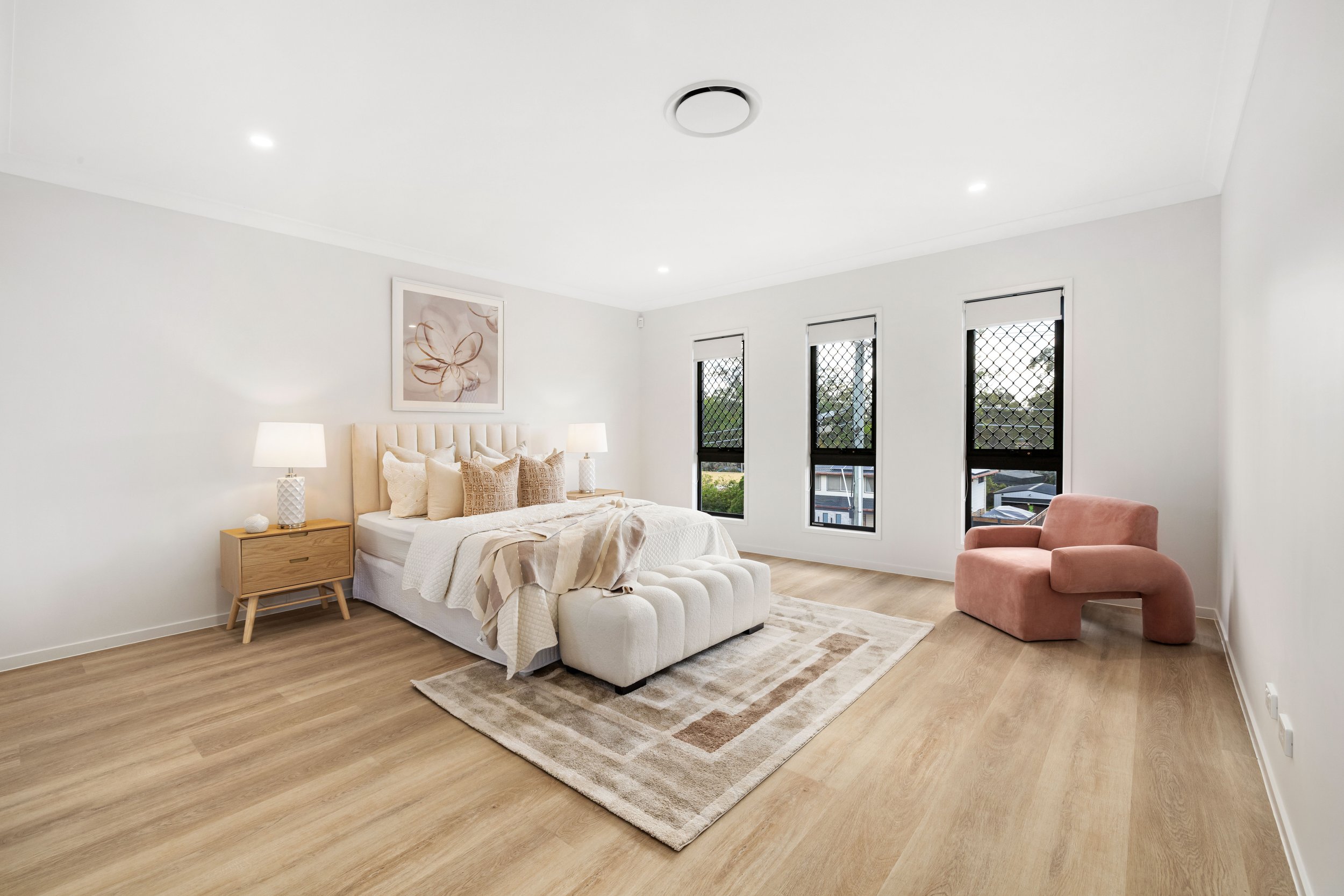 A spacious bedroom with a large bed, beige upholstered headboard, multiple pillows, bedside tables with lamps, a pink curved armchair, and three windows with black frames and security screens, wooden flooring, and a light-colored area rug.