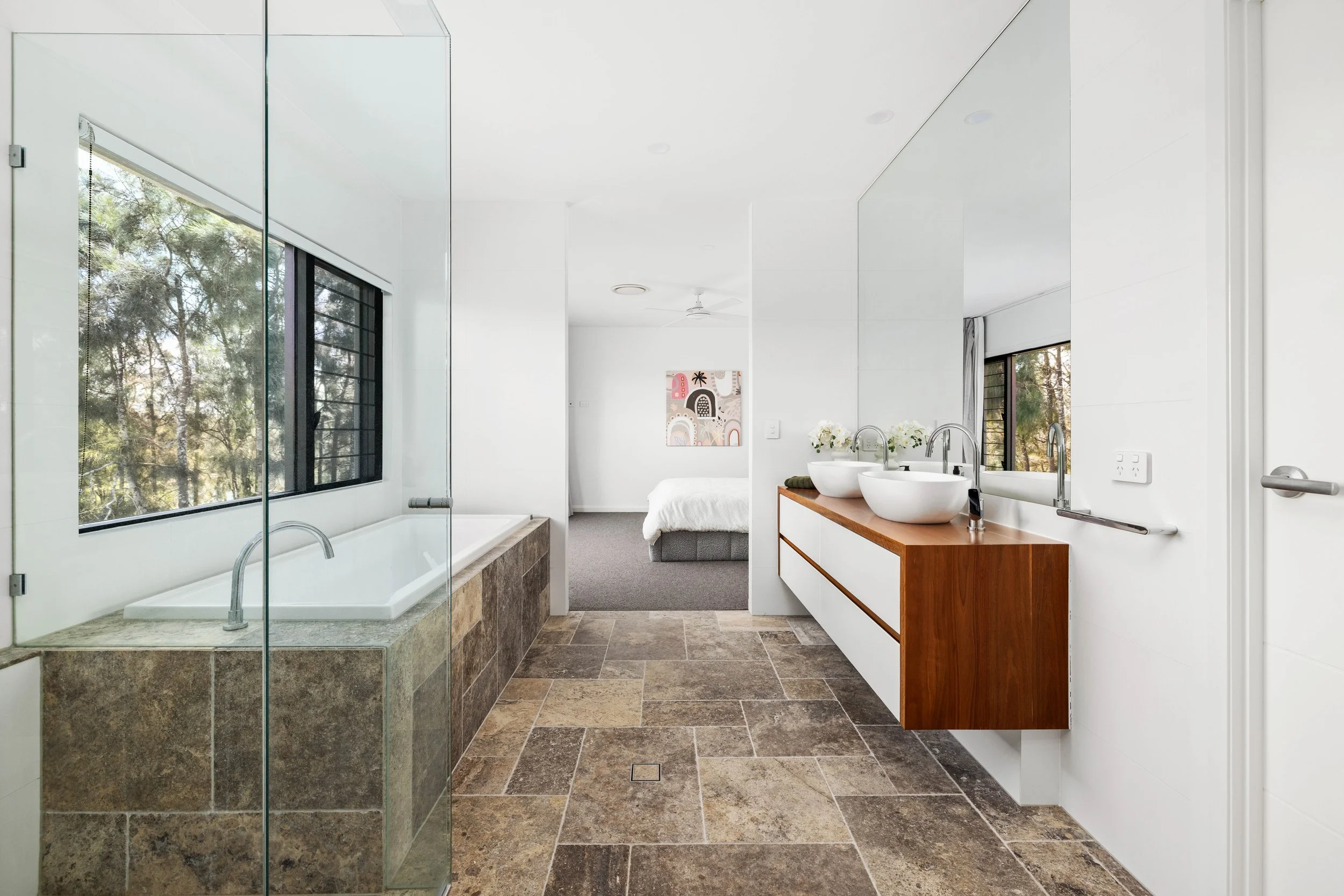Modern bathroom with a bathtub next to a window, a wooden vanity with two vessel sinks, and a mirror reflecting the rest of the room, with a bedroom in the background.