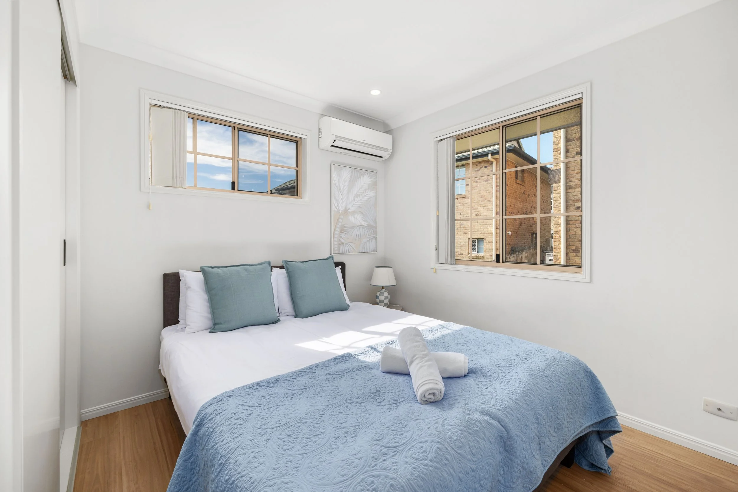 Bright bedroom with white walls, a bed with a blue quilt and three teal pillows, two rolled towels on the bed, a nightstand with a lamp, large windows showing neighboring brick buildings, and an air conditioning unit.