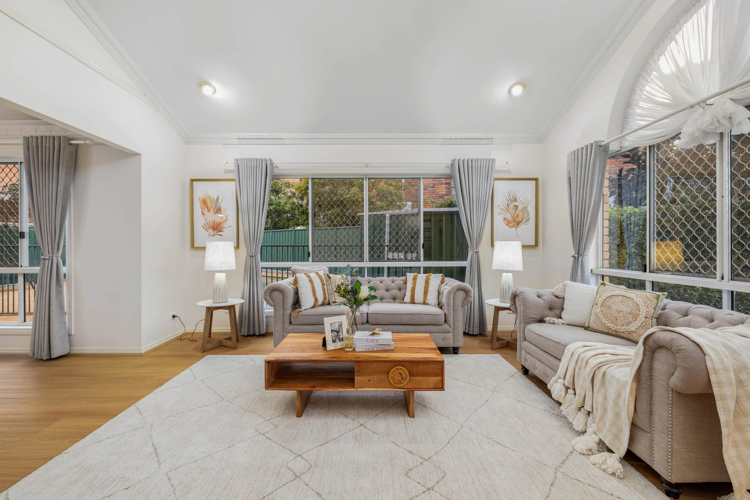 Bright living room with white walls, light grey sofas, wooden coffee table, and large windows with grey curtains, in a cozy modern style.