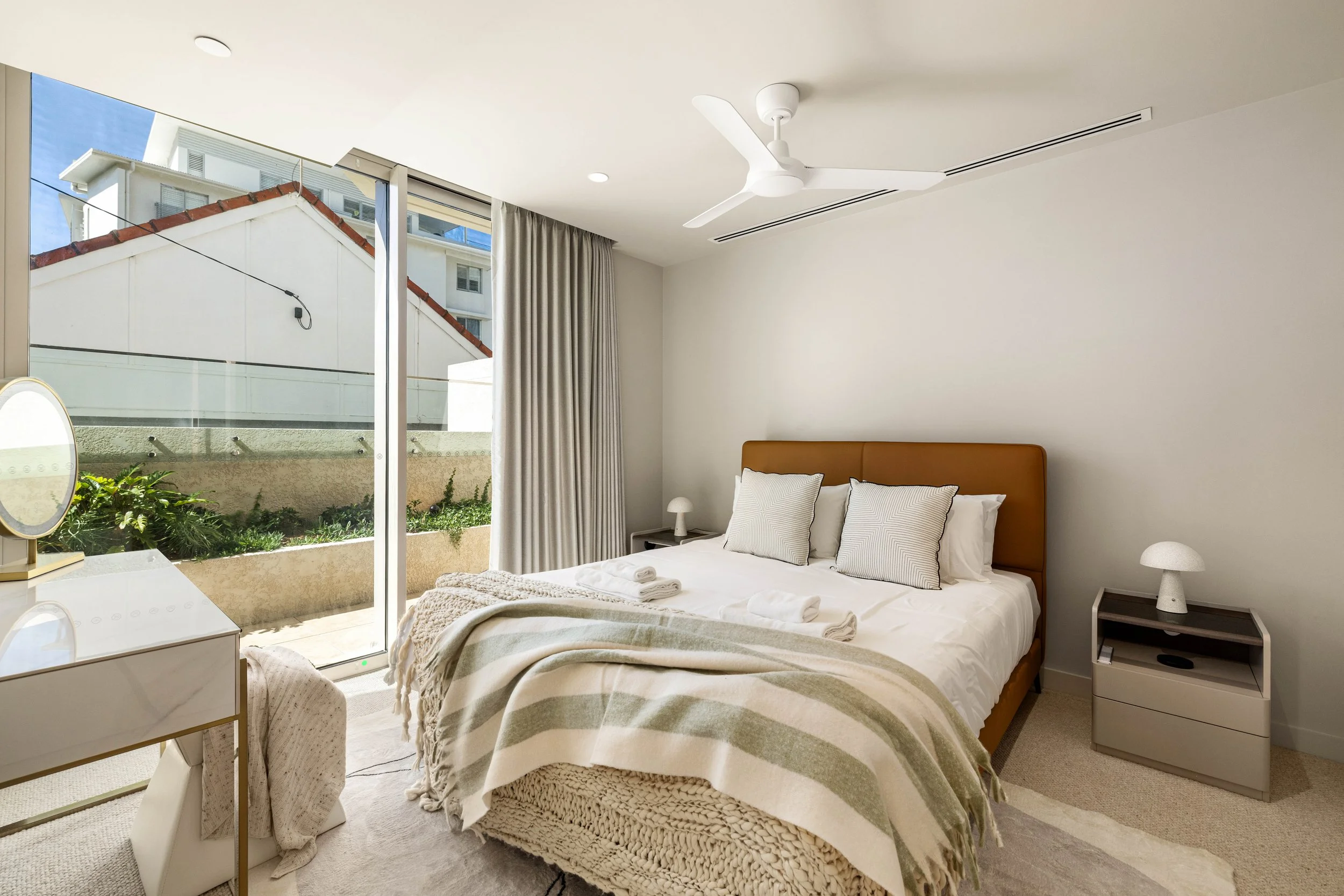 Modern bedroom with a large bed, neutral tones, and natural light from a glass sliding door leading to a small outdoor garden.
