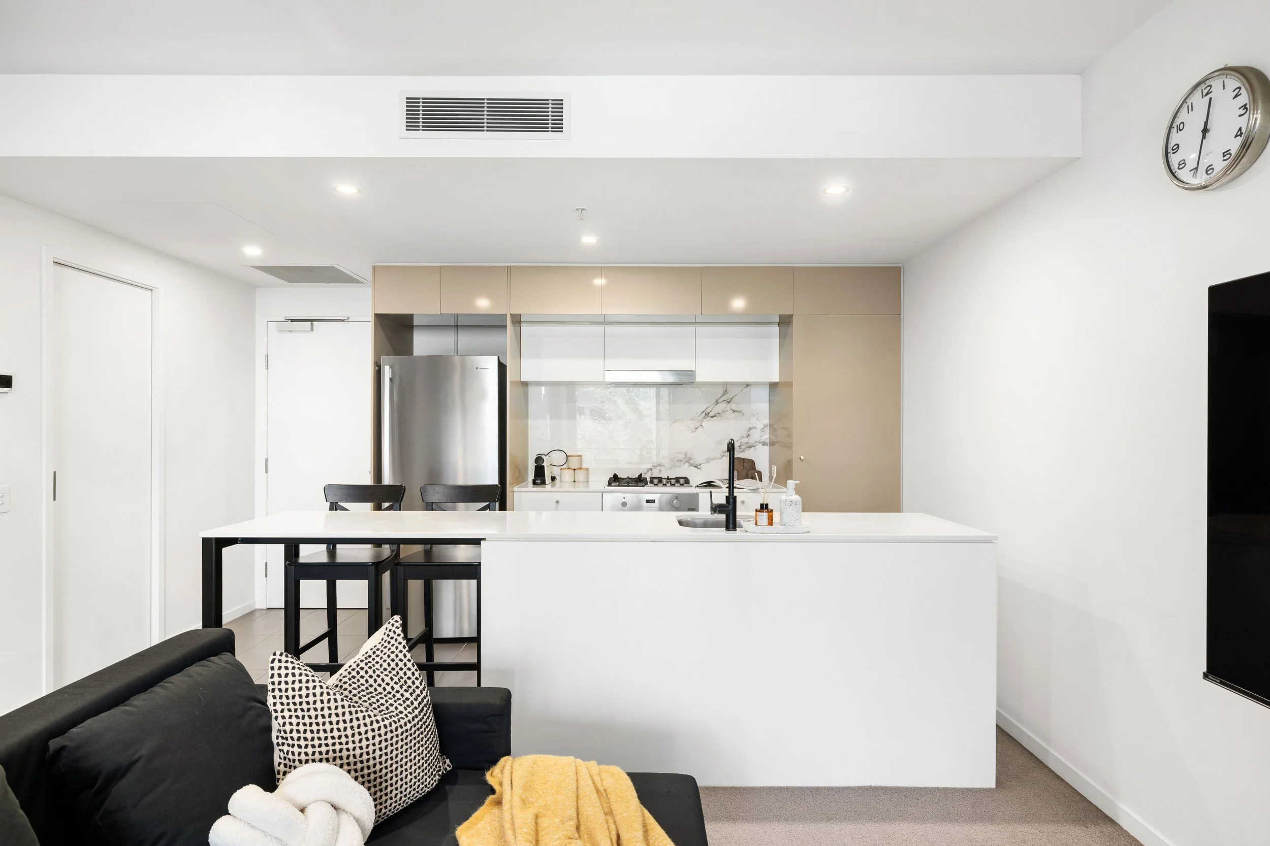 Modern kitchen in an apartment with white countertops, beige cabinets, stainless steel refrigerator, marble backsplash, black faucet, and a small breakfast bar with two black chairs. Part of a black sofa with patterned pillows and a yellow blanket vi