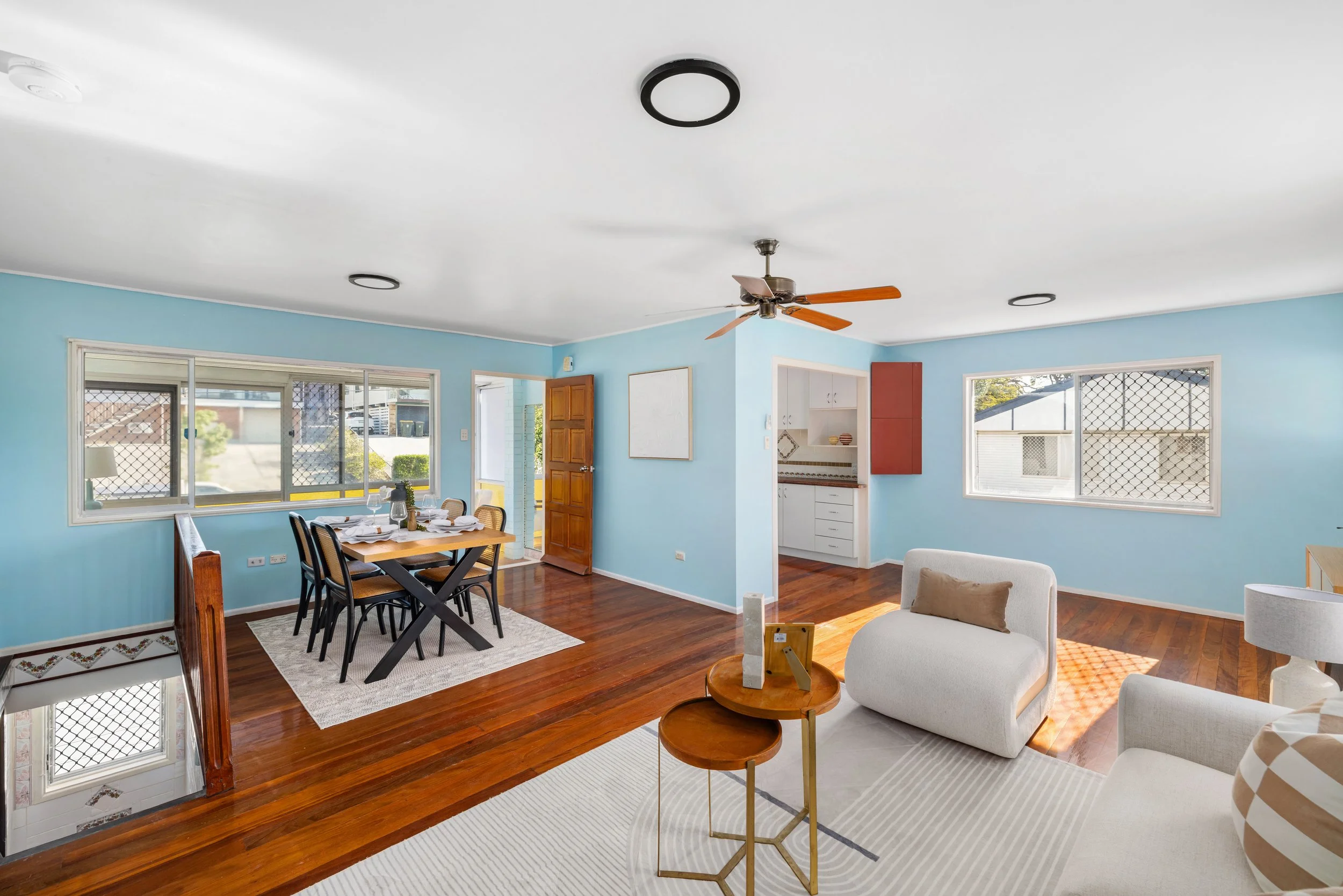 Living room with light blue walls, hardwood floors, a white couch, a white armchair with a beige pillow, and a small round wooden table. The room has large windows with security screens and a ceiling fan with wooden blades. In the background, there i