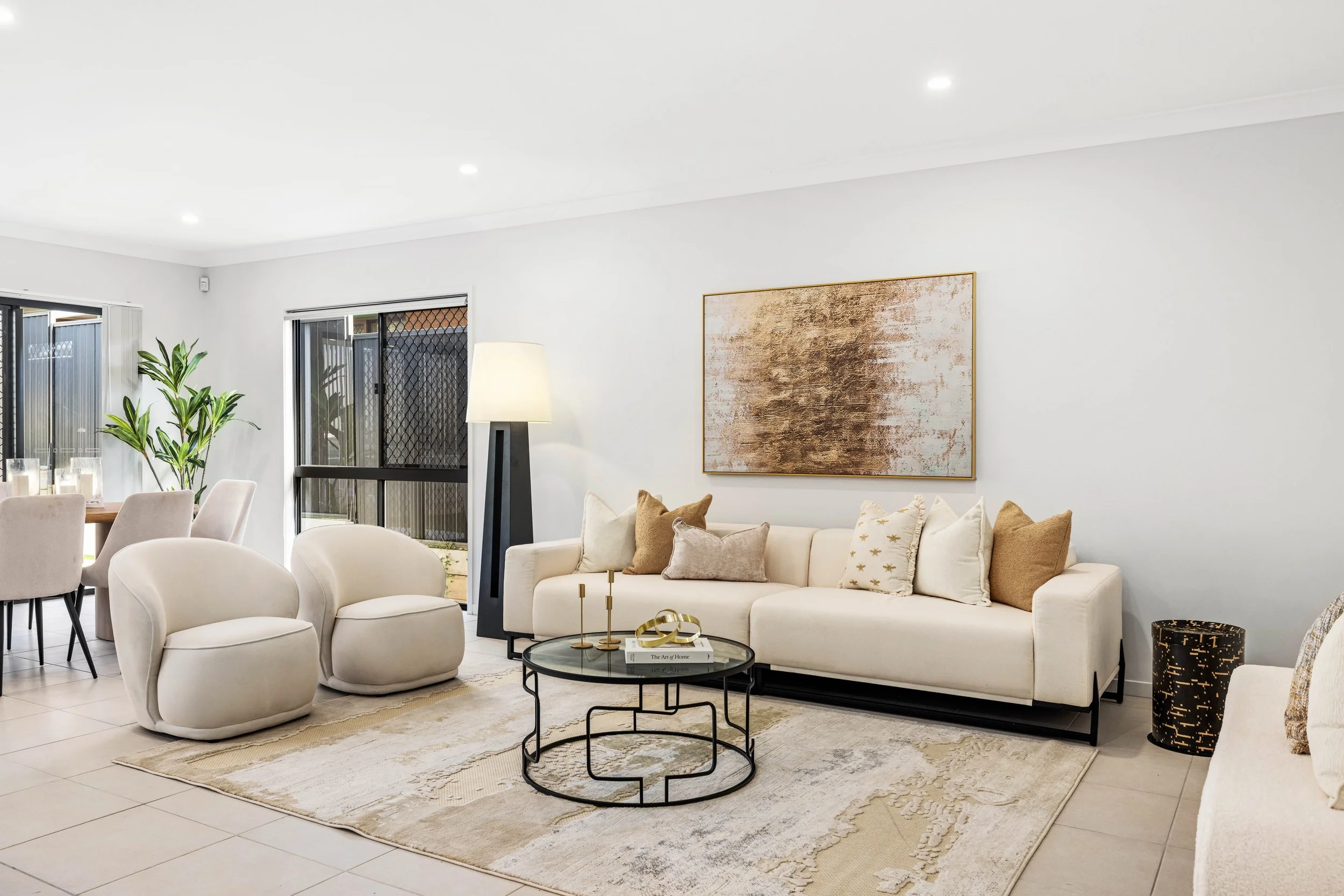 Modern living room with white sofa, beige and gold throw pillows, glass coffee table, abstract wall art, tall floor lamp, beige arching chairs, potted plant, and large windows.