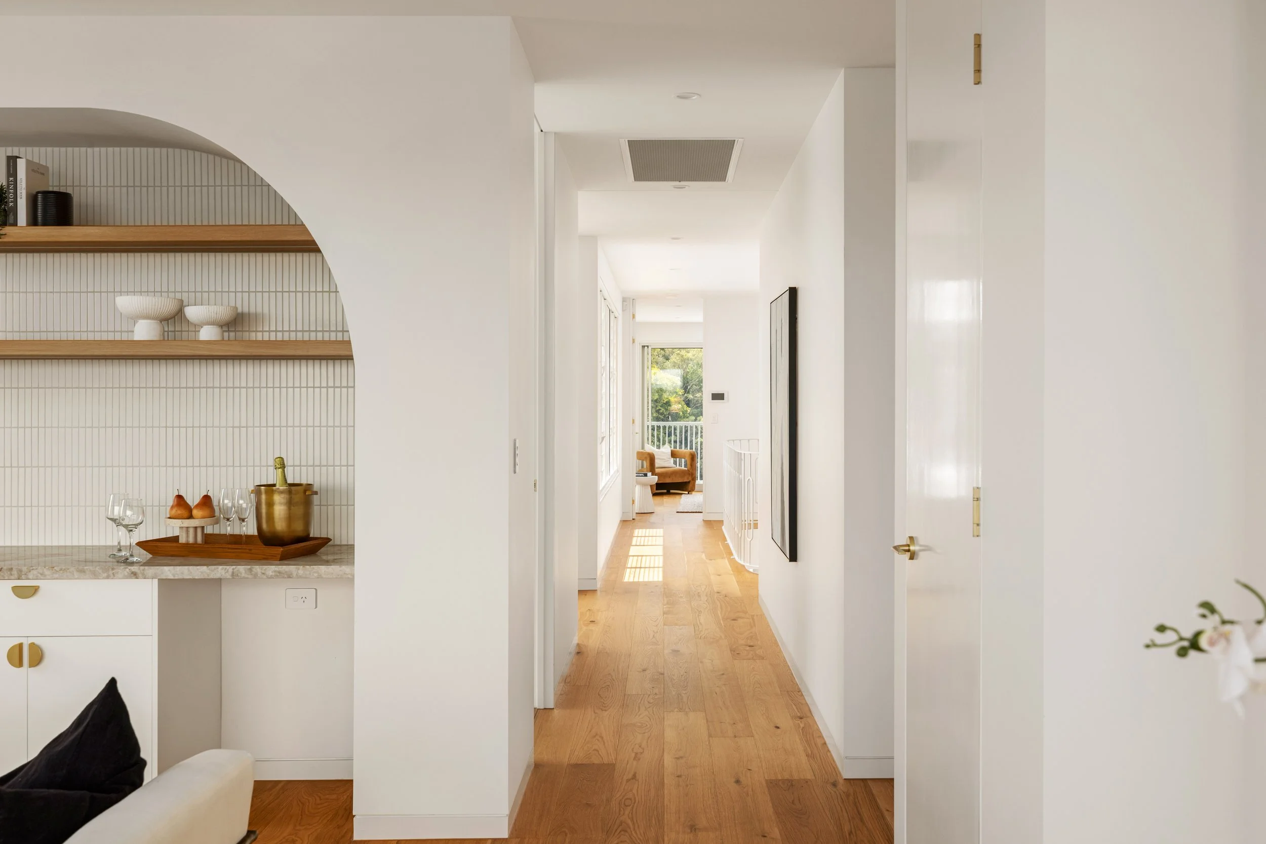 Bright hallway with wooden flooring, leading to a sunlit room with outdoor balcony view, white walls, and minimal decor.