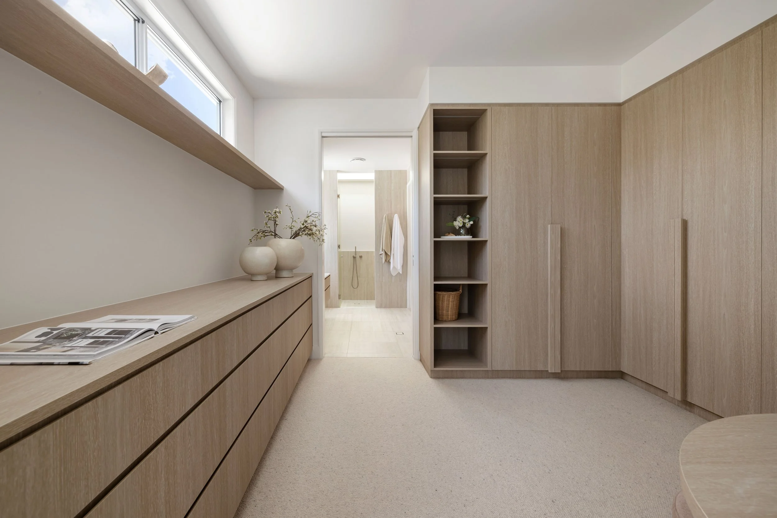 Minimalist bedroom with light wood furniture, including a long dresser with decorative vases, open shelves with flowers and a basket, built-in wardrobes, a window with a ledge, and a view into a bathroom with towels.
