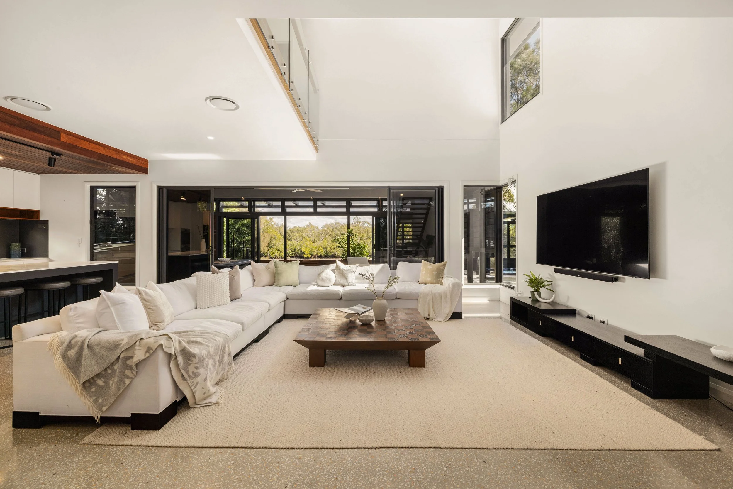 Modern living room with large white sectional sofa, wooden coffee table, black wall-mounted TV, and floor-to-ceiling windows showing outdoor greenery.