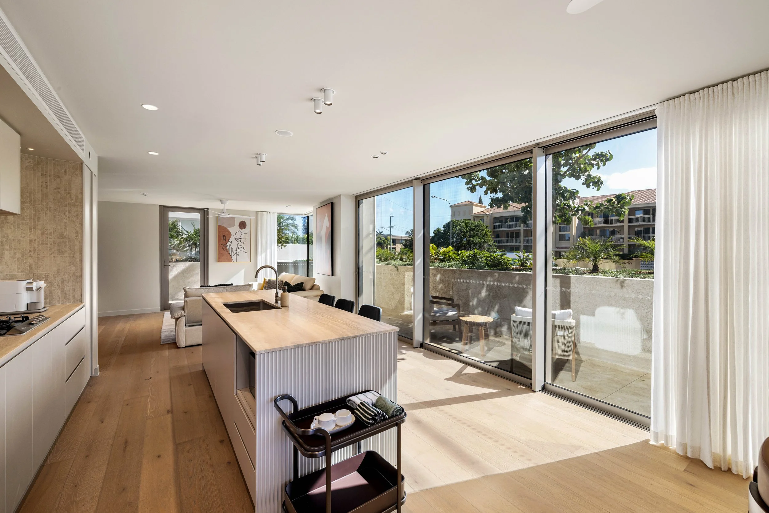 Bright, open-concept living room and kitchen with large glass sliding doors leading to a balcony with outdoor seating, view of trees and apartment buildings outside, modern decor, white curtains, wooden flooring.