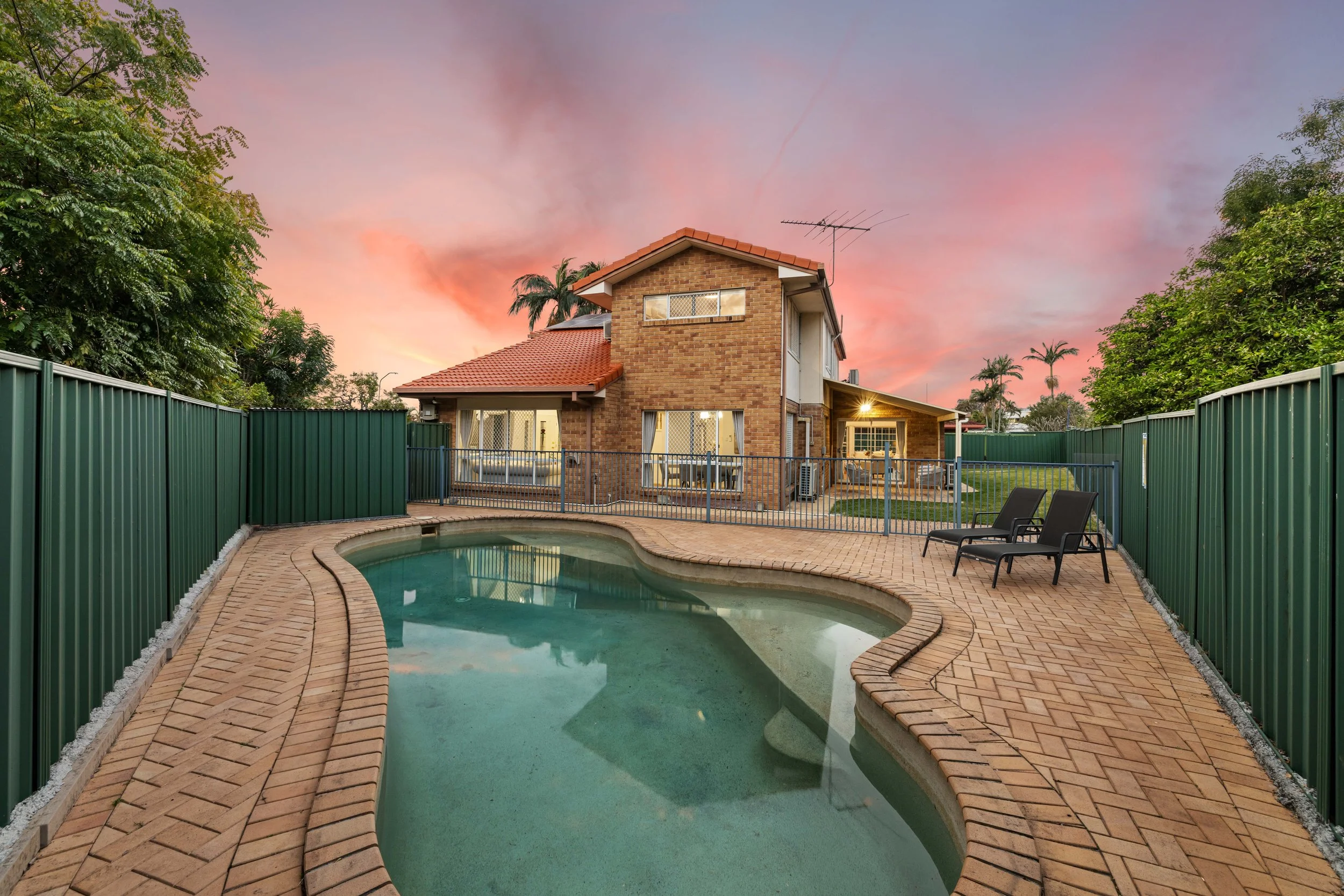 Backyard with a kidney-shaped swimming pool, surrounded by brick paving, enclosed by green metal fencing, and a two-story brick house with large windows and a patio, under a pink and orange sunset sky with some trees in the background.