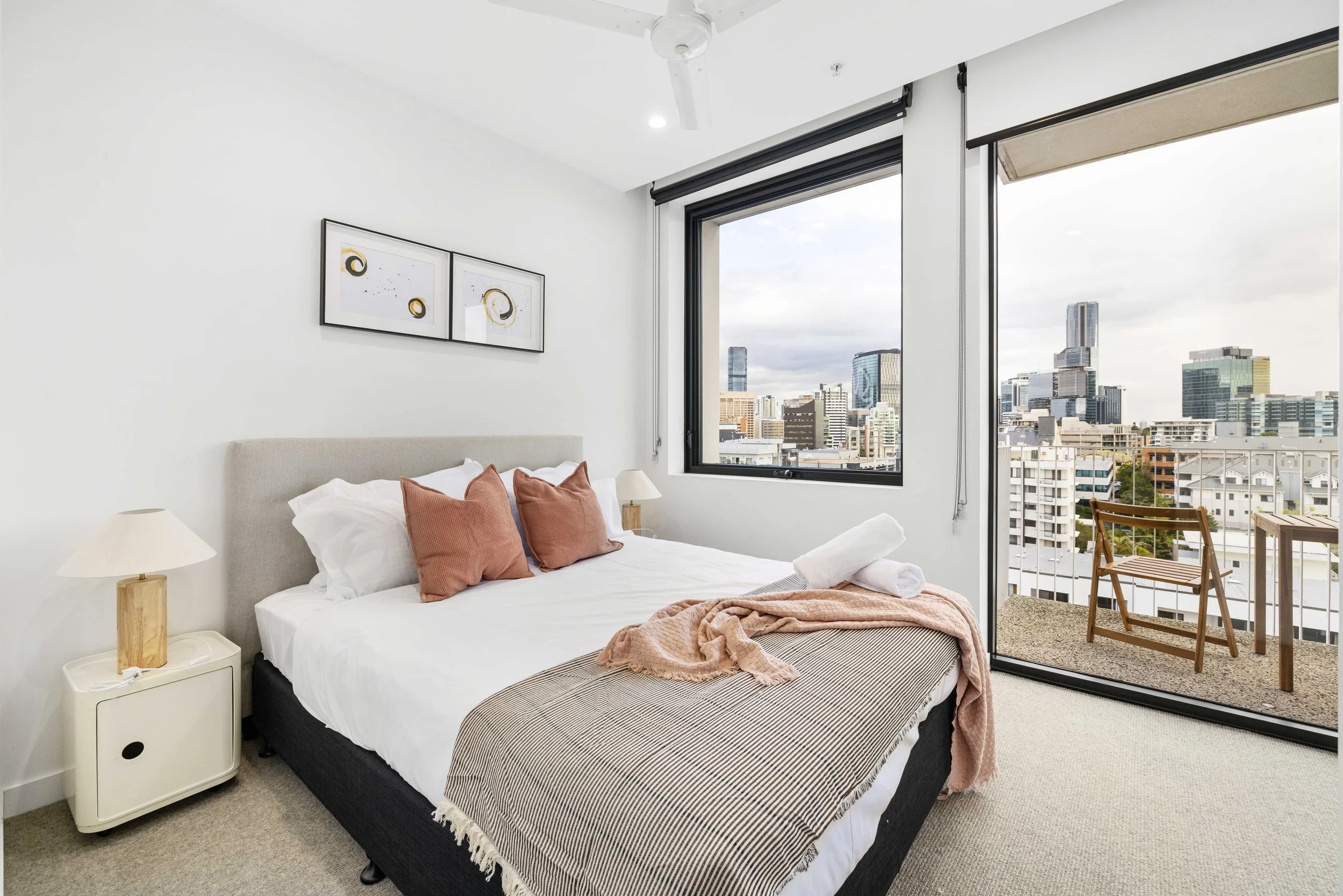 Modern bedroom with large window overlooking city skyline, bed with white bedding and brown pillows, small table with lamp, balcony with wooden chair and view of high-rise buildings.