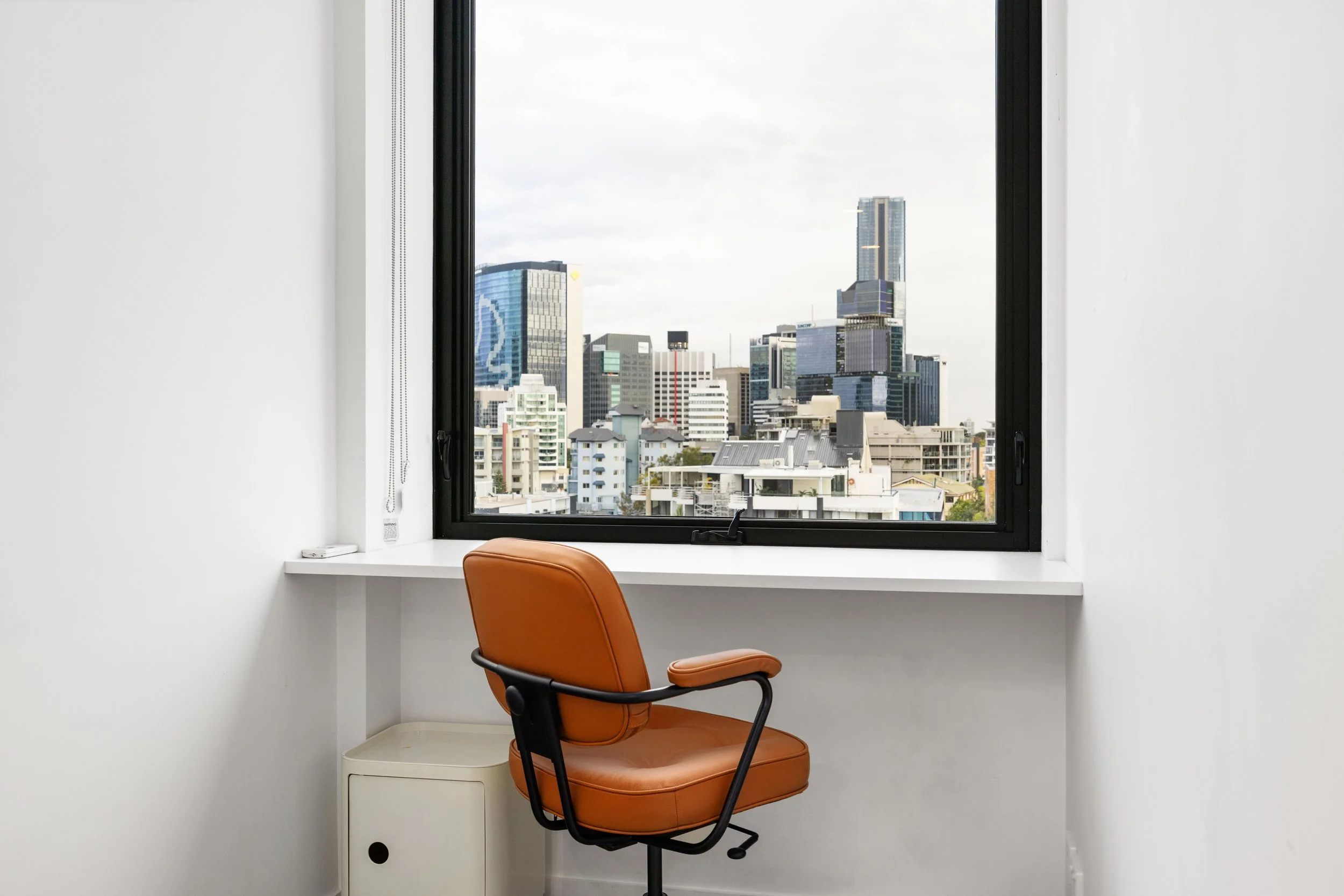 An office window view with a cityscape of tall buildings, a brown office chair, and a white desk or ledge.