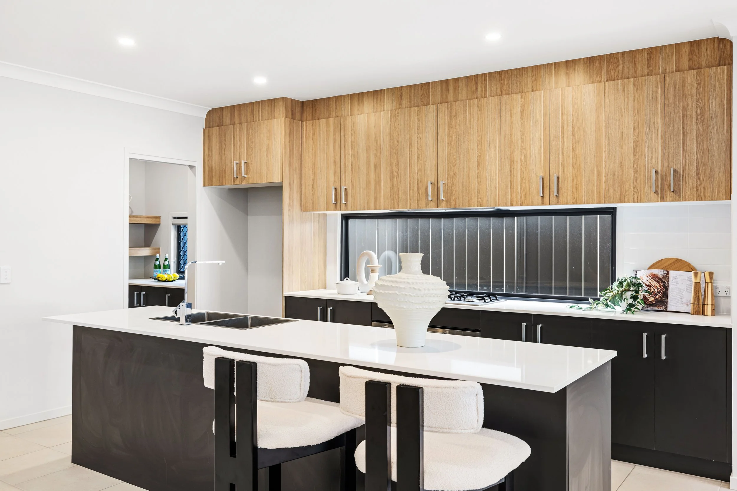 Modern kitchen with black and wooden cabinets, white countertops, and a white island with two bar stools. Decor includes a large white vase, a small white pitcher, a book, and some greenery on the counter.