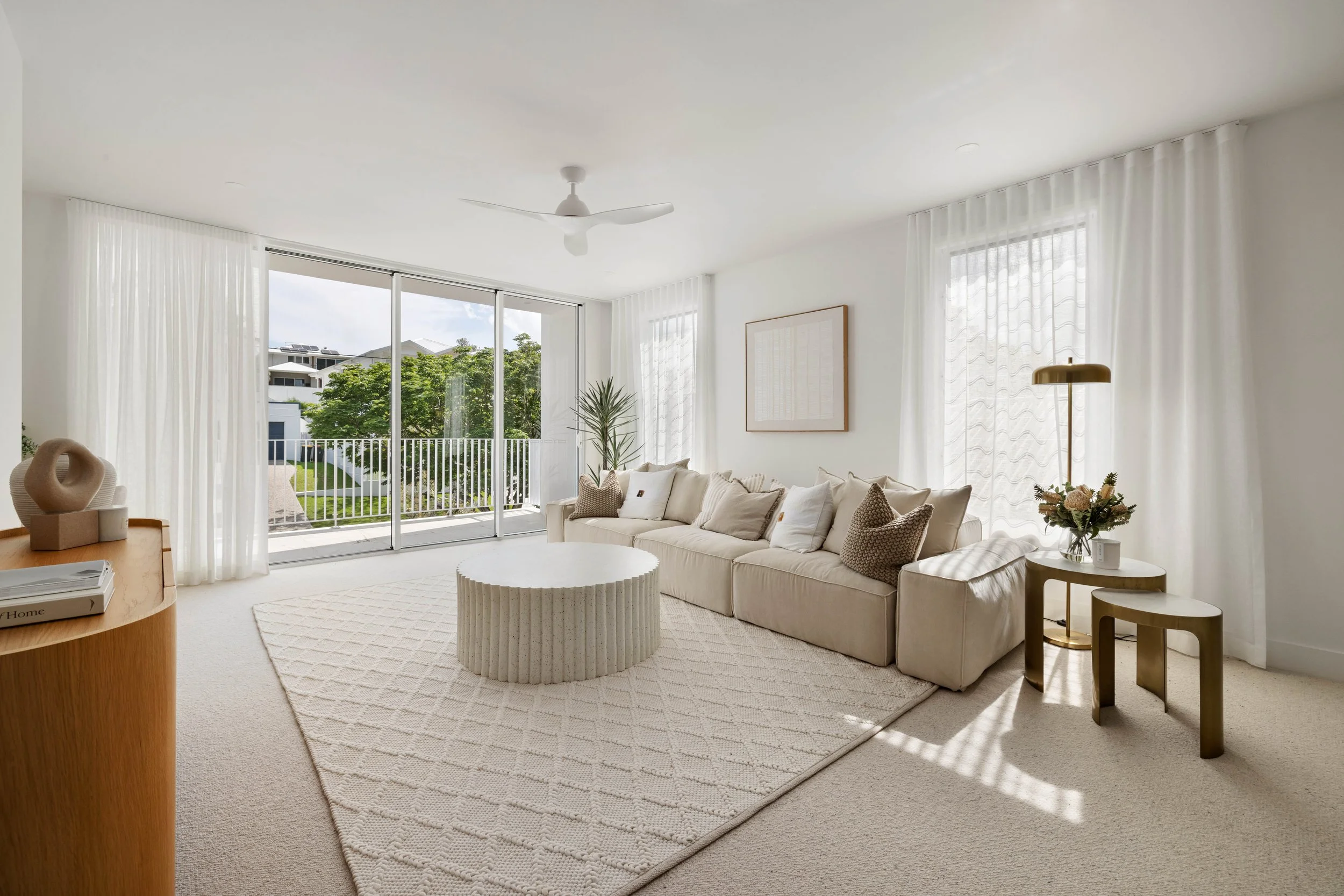 Bright living room with large sliding glass doors opening to a balcony, white walls, a beige sectional sofa with multiple pillows, a patterned rug, small side tables with a flower vase and lamp, a ceiling fan, and sheer white curtains.