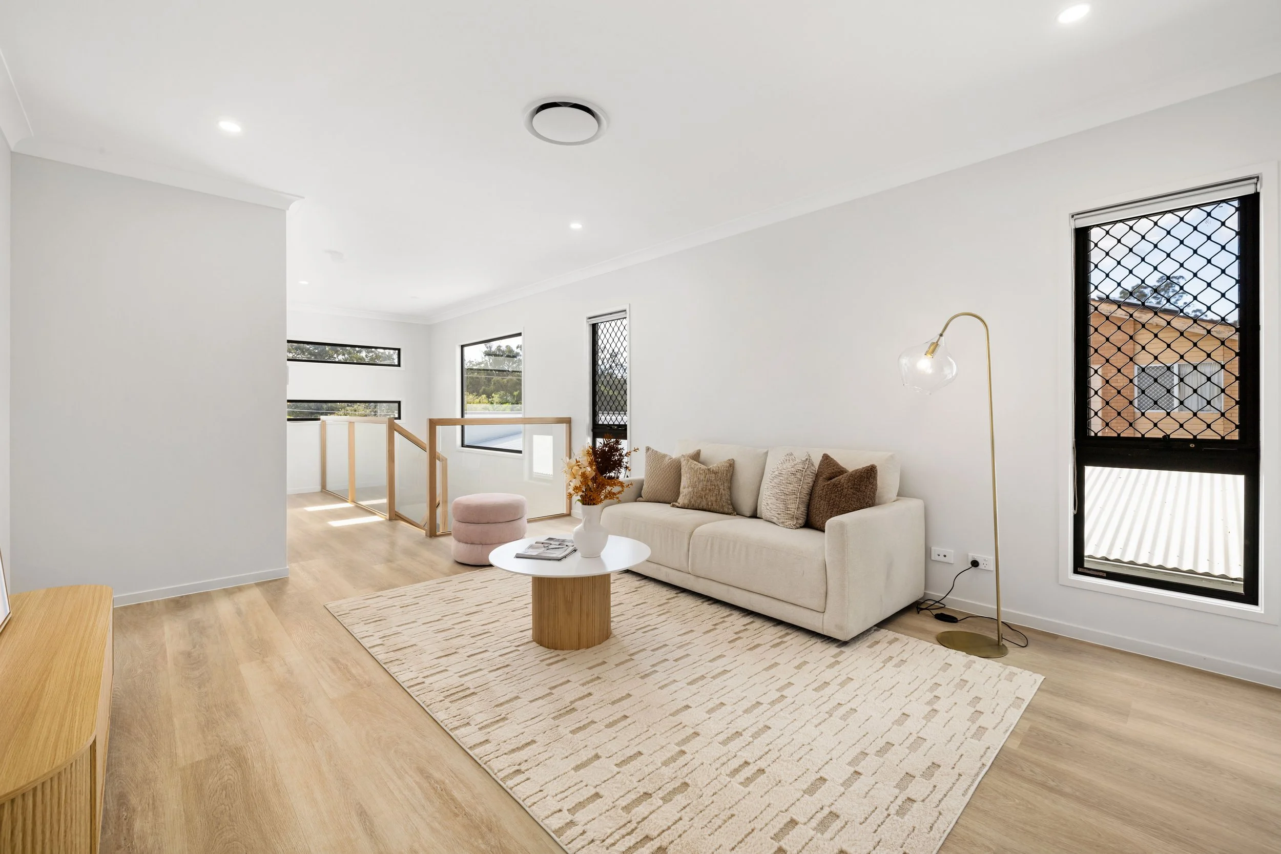 Bright living room with white sofa, beige cushions, wooden coffee table with flowers, pink ottoman, area rug, and large black-framed windows with security screens, light wooden floor, and modern floor lamp.