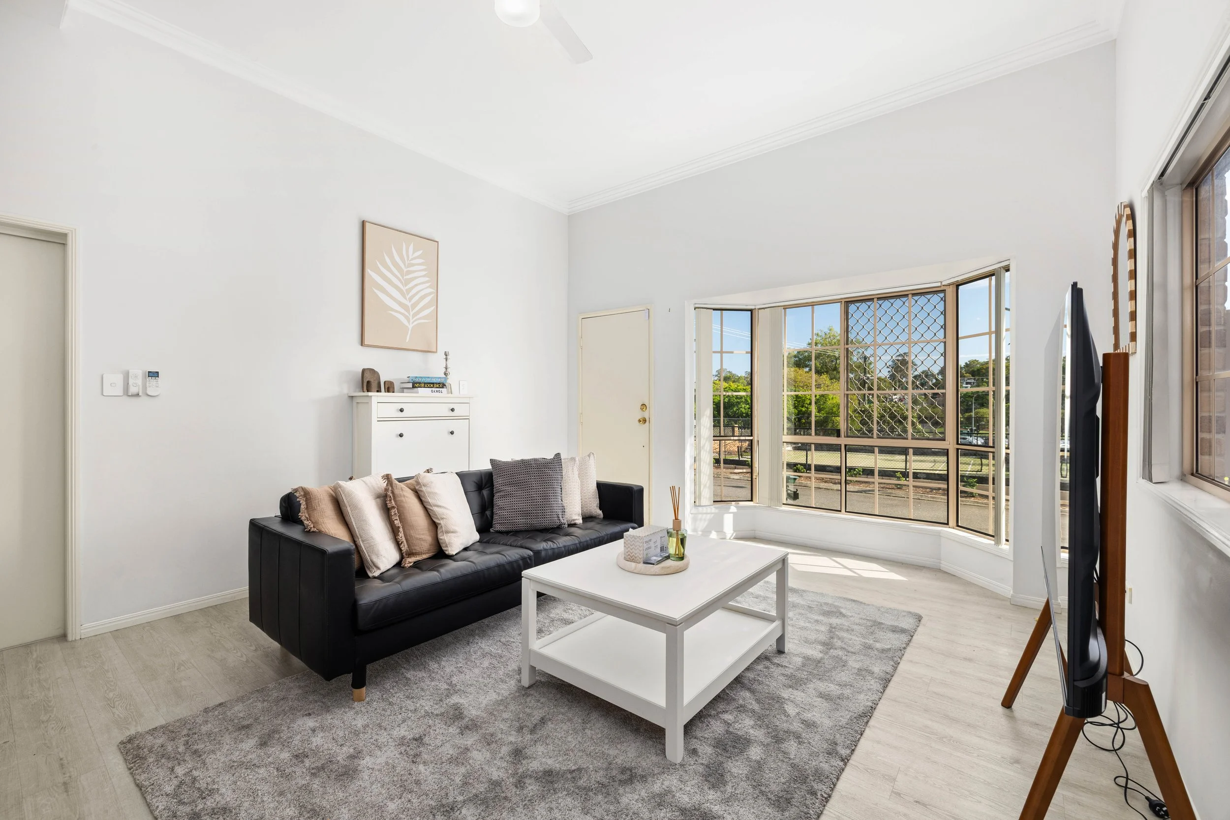 Bright living room with white walls, black sofa with beige and gray pillows, white coffee table, large bay window with grid pattern, TV on wooden stand, gray area rug, and decorative artwork on wall.