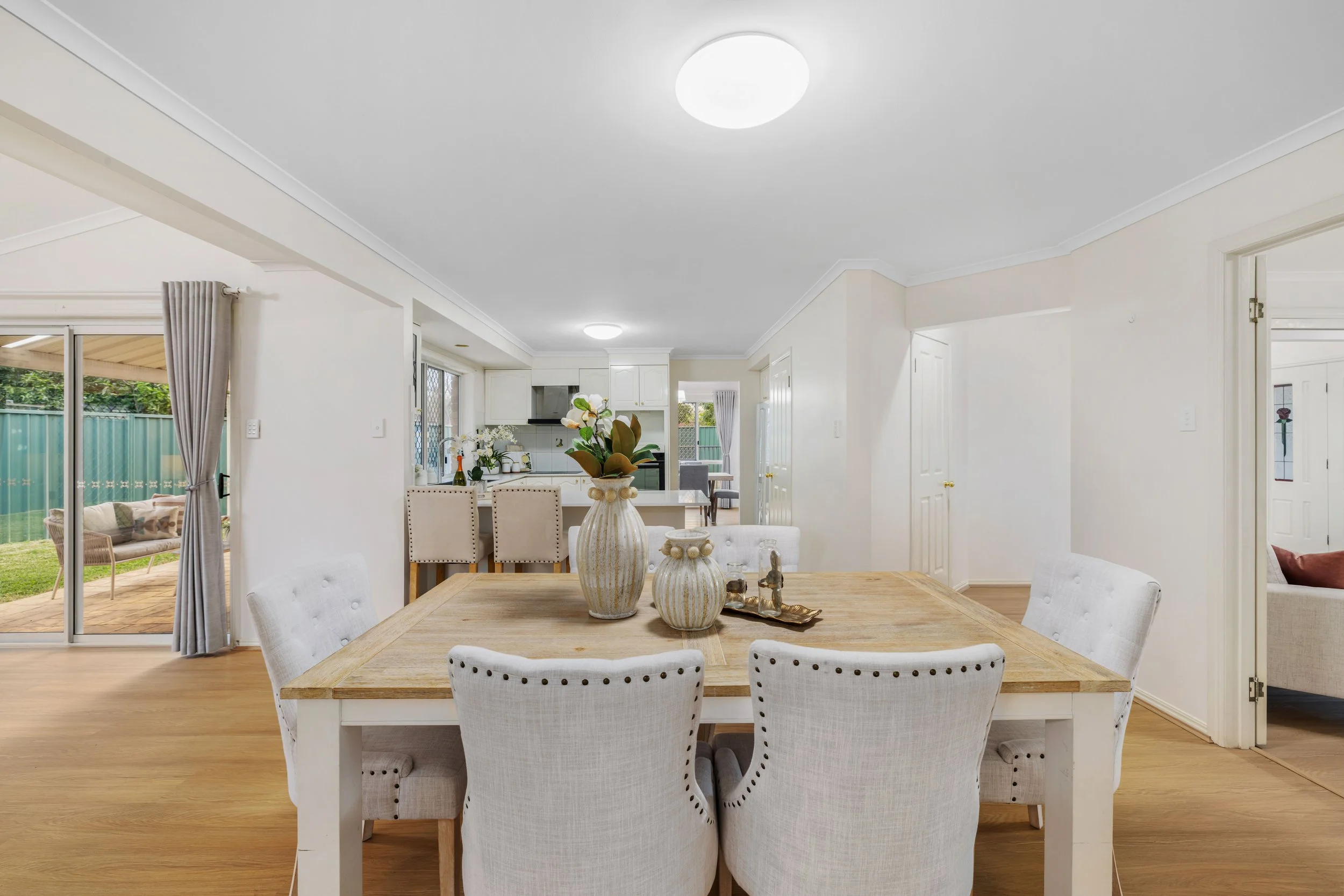 A dining room with a wooden table, six white upholstered chairs, and a centerpiece of vases with flowers on the table. In the background, an open kitchen with bar stools, a sliding glass door to the outside patio, and a view into a living area.