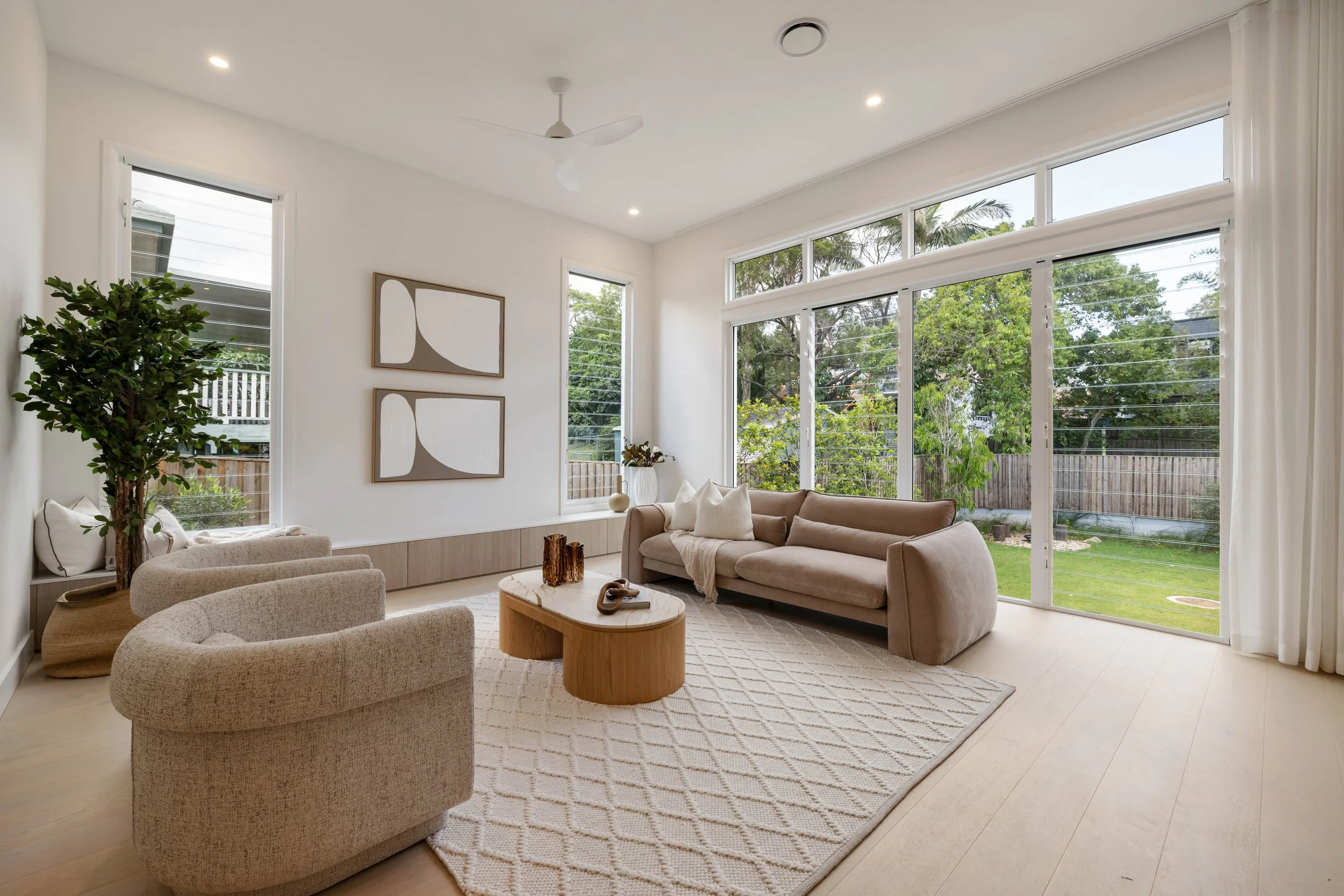 Bright living room with large windows, beige sofa, armchair, wooden coffee table, potted plant, and modern wall art.
