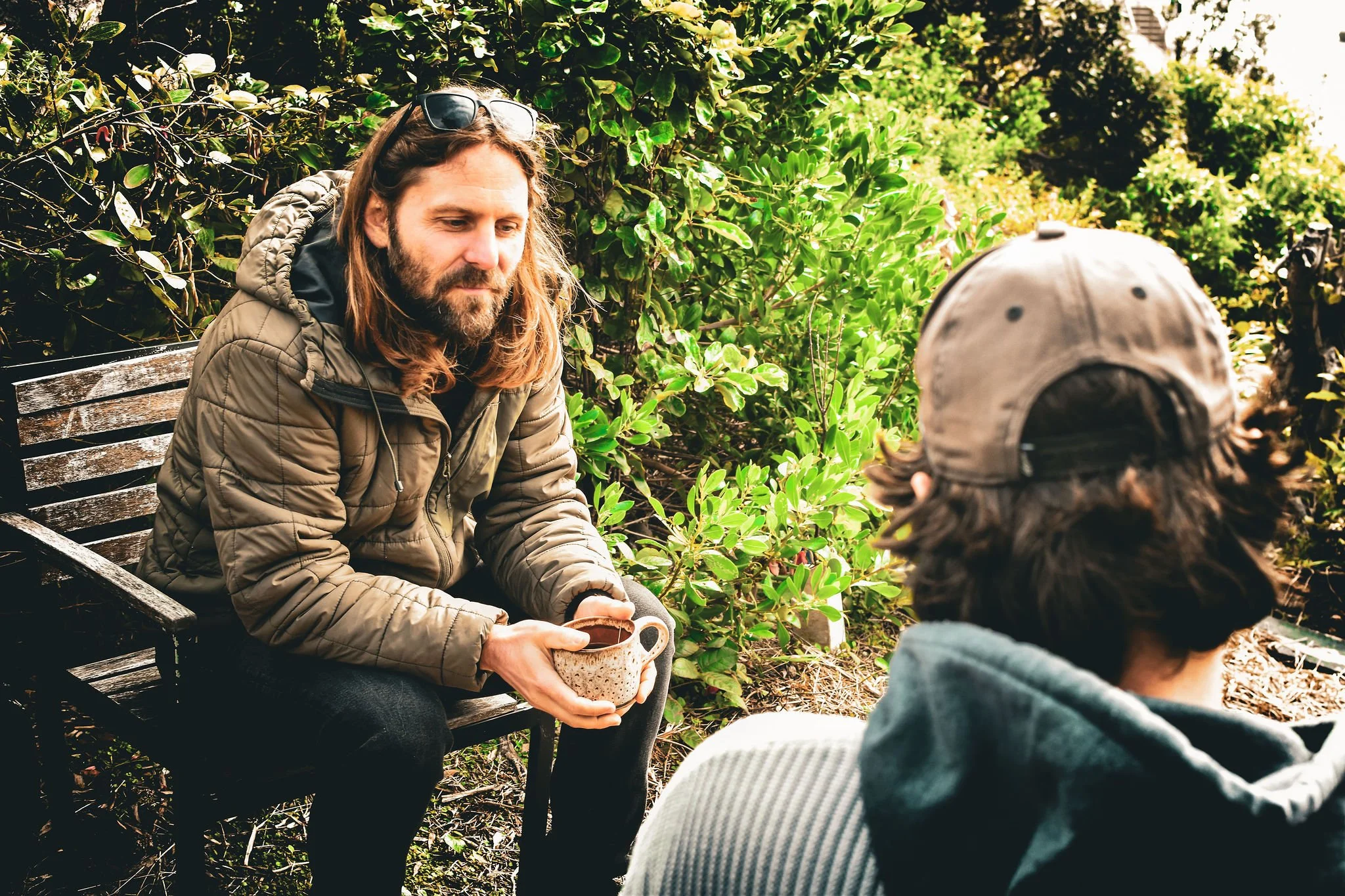 Two men sitting outside in a garden, one with long hair and beard, holding a mug, the other with a cap and hoodie, facing each other among lush green bushes.