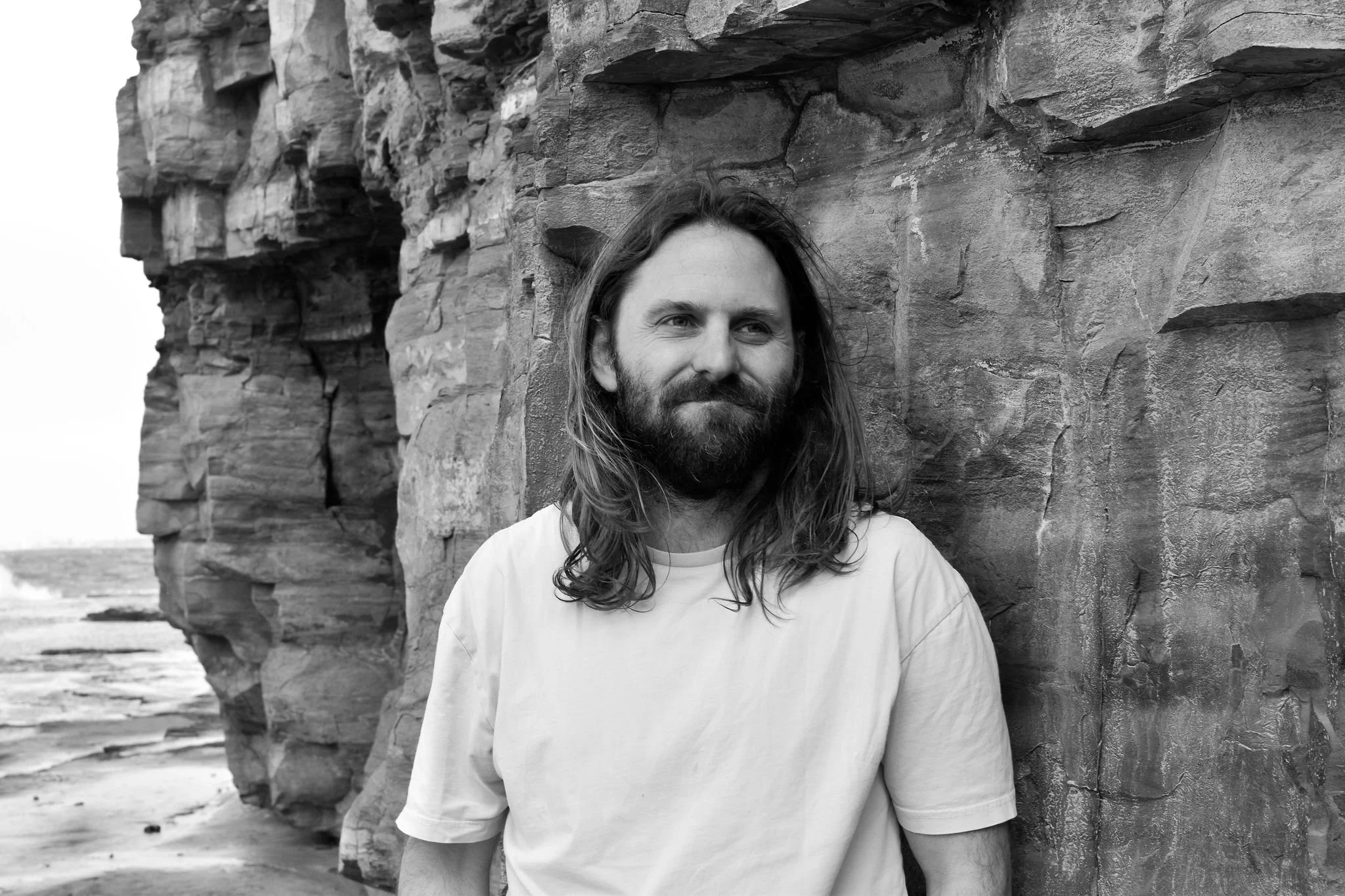 A man with long hair and a beard standing against a rocky cliff on a beach.