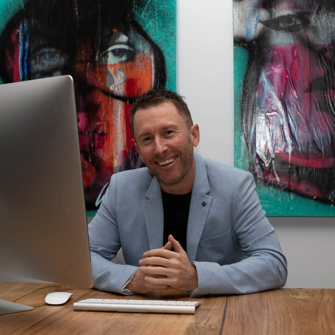 A man with short brown hair, beard, and dressed in a light gray blazer and black shirt, sitting at a wooden desk with a large computer monitor, mouse, and keyboard, smiling at the camera. Behind him are colorful abstract portrait paintings of faces.