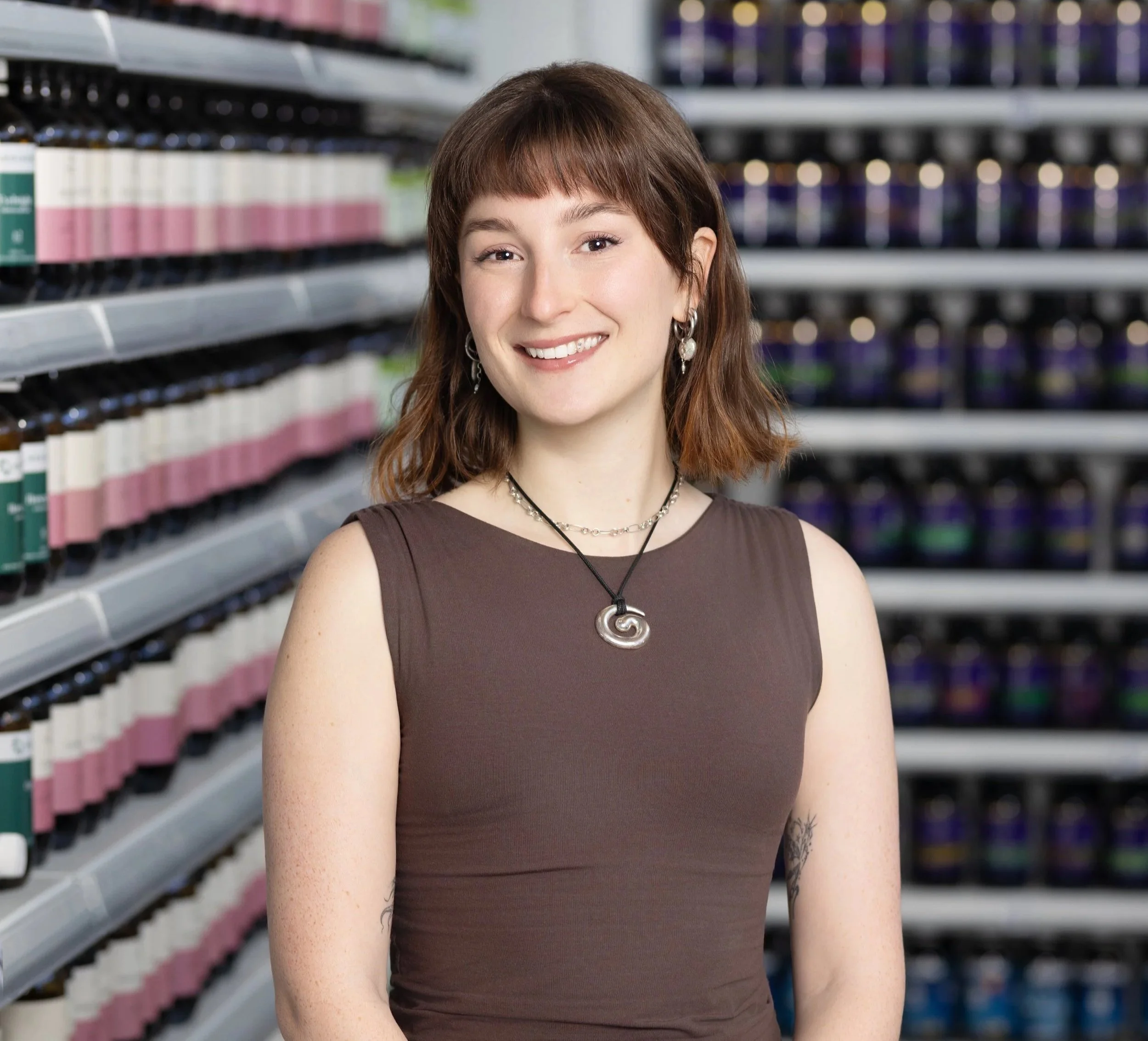 A young woman with brown hair and bangs standing in a store aisle with shelves of products in the background, smiling at the camera.