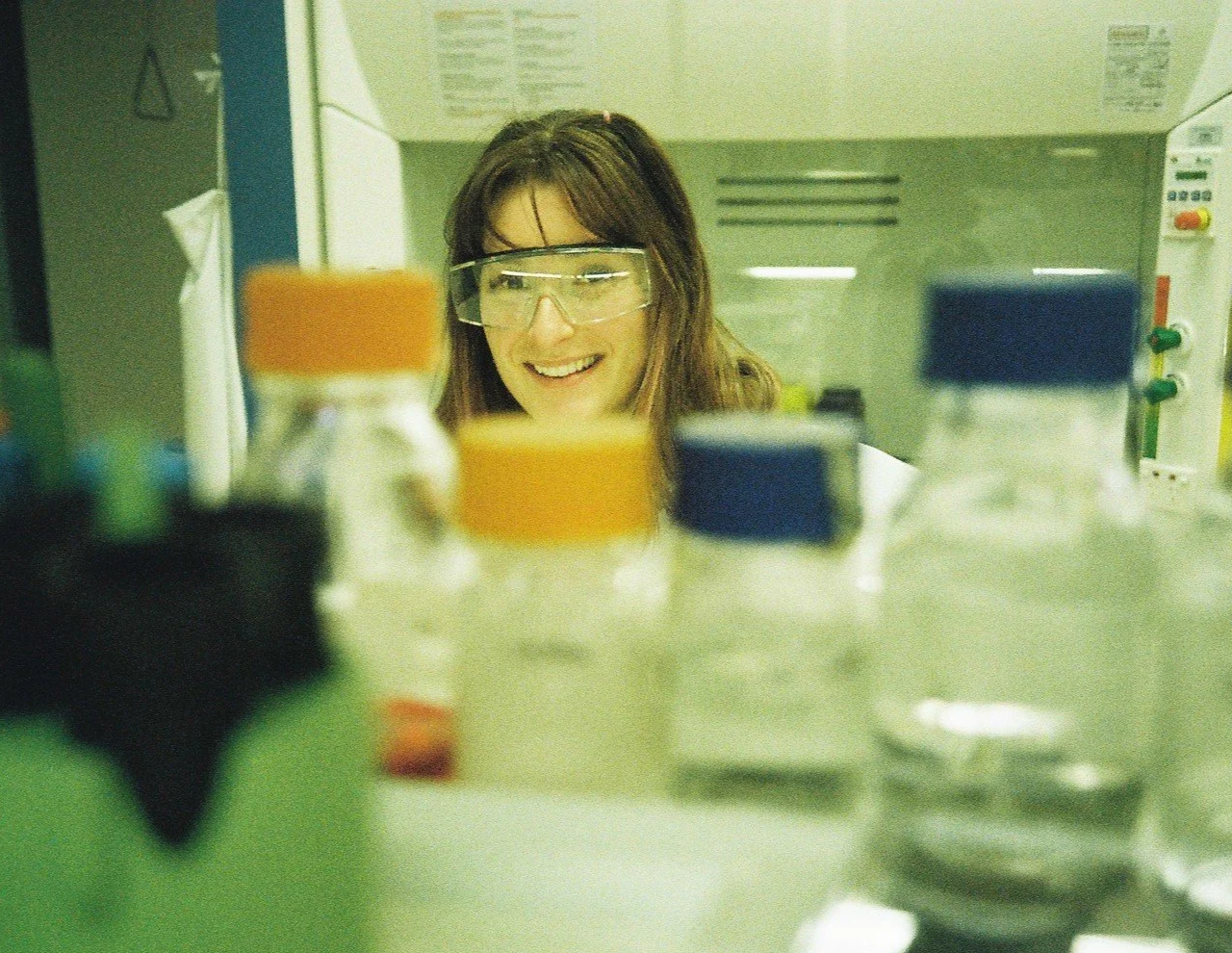 A woman wearing safety goggles smiling as she looks at laboratory bottles filled with liquids, placed on a lab bench.