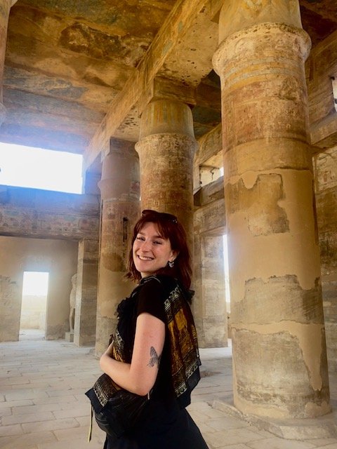 A young woman with red hair smiling inside a historic stone structure with large columns and high ceilings.