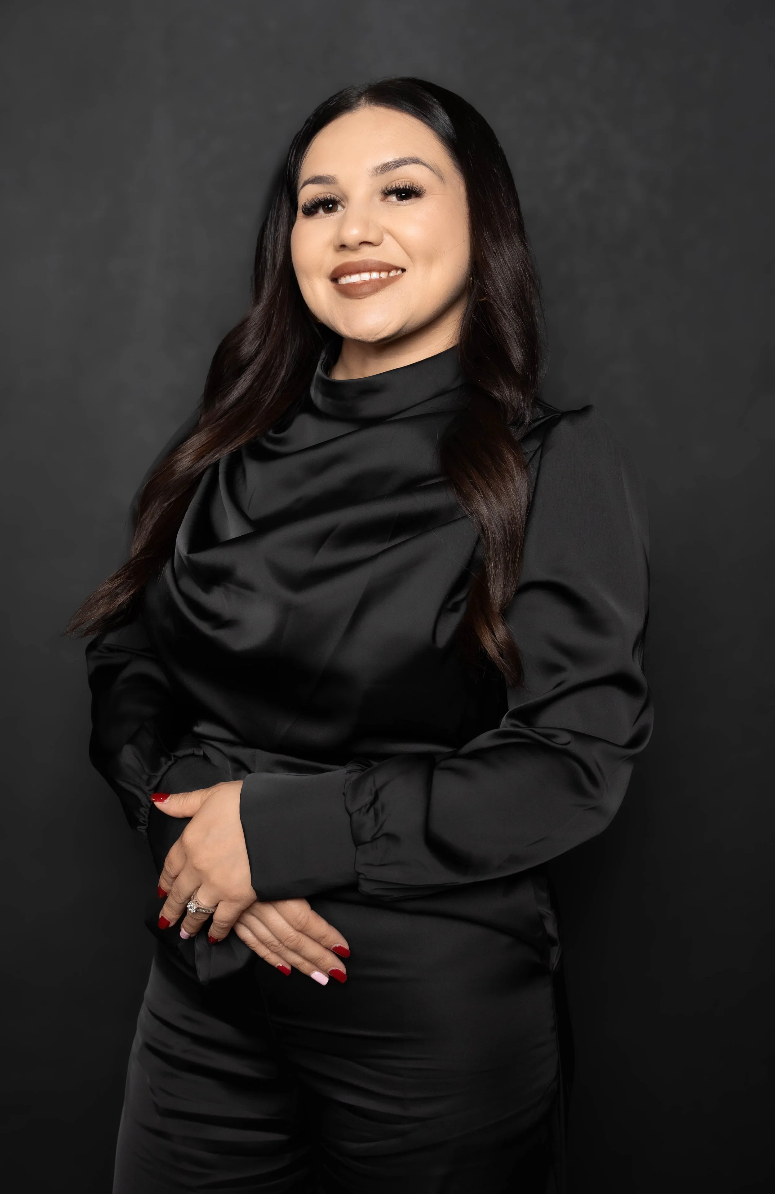 A woman with long dark hair wearing a black satin blouse and matching black pants, standing against a dark background.