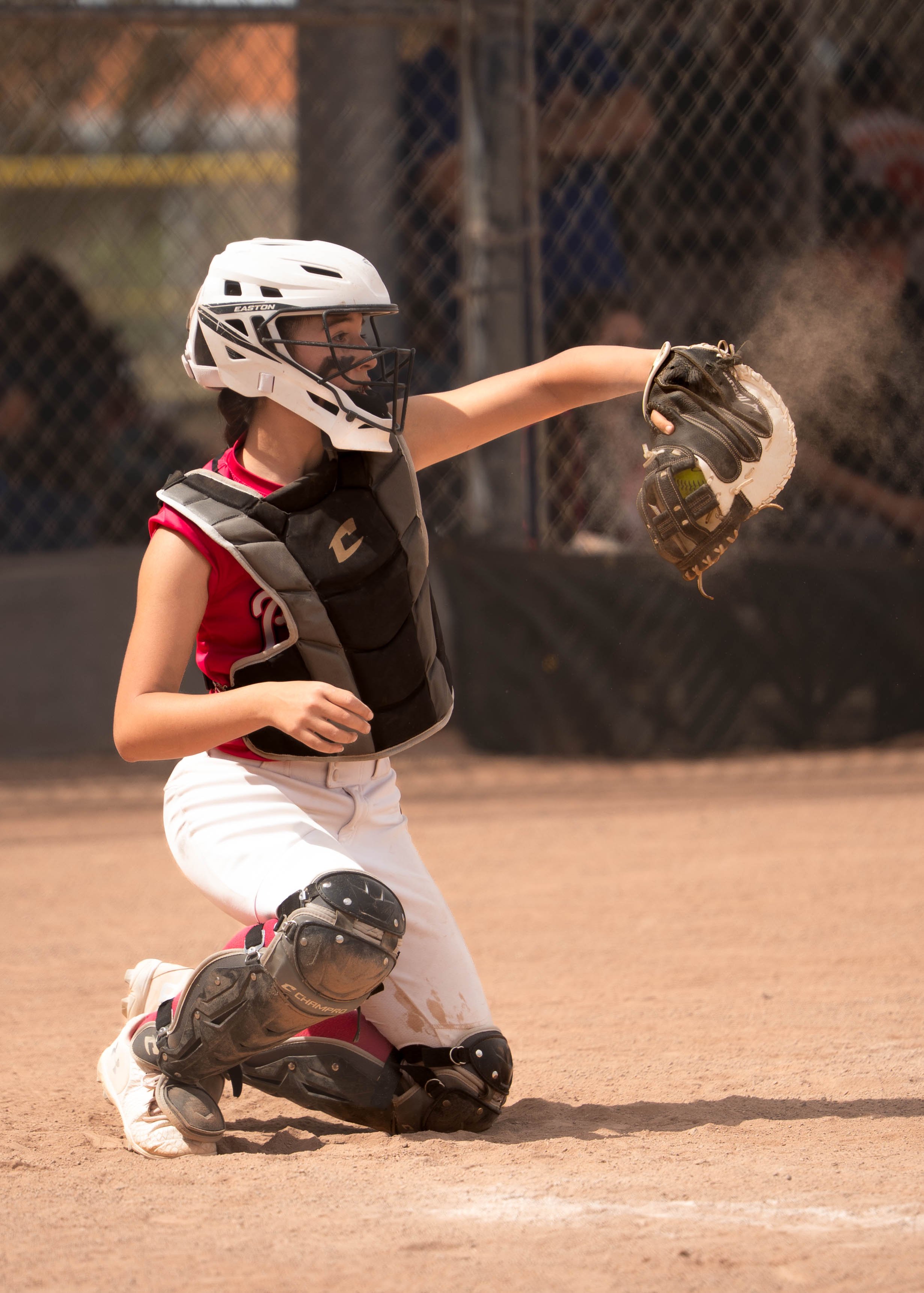 A female baseball catcher wearing a helmet, protective gear, and a glove, kneeling on the dirt field during a game, making a throw.