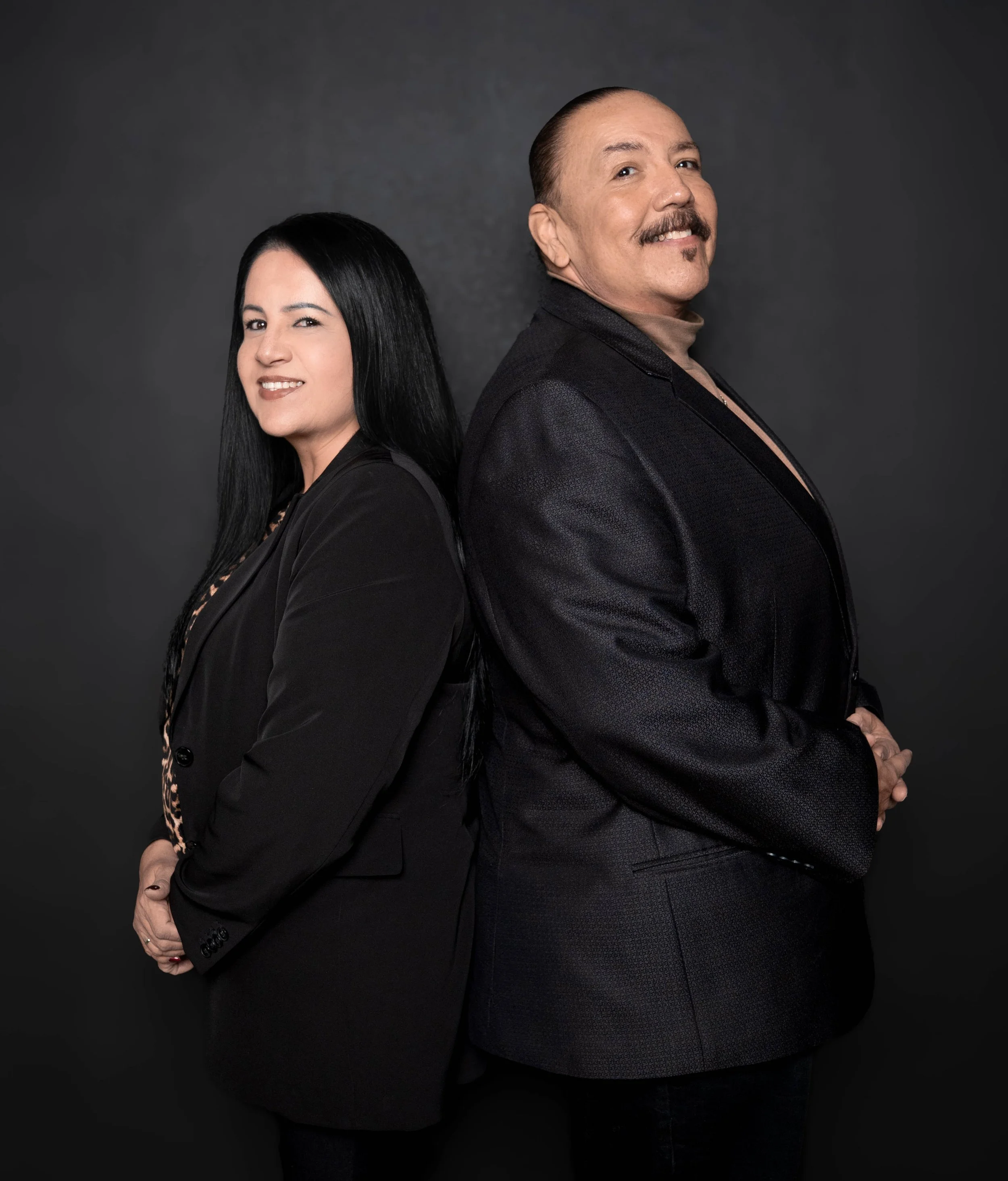 A woman with long black hair and a man with a mustache and goatee stand back-to-back against a dark background, both dressed in black suits. The woman is smiling slightly, and the man is smiling broadly.