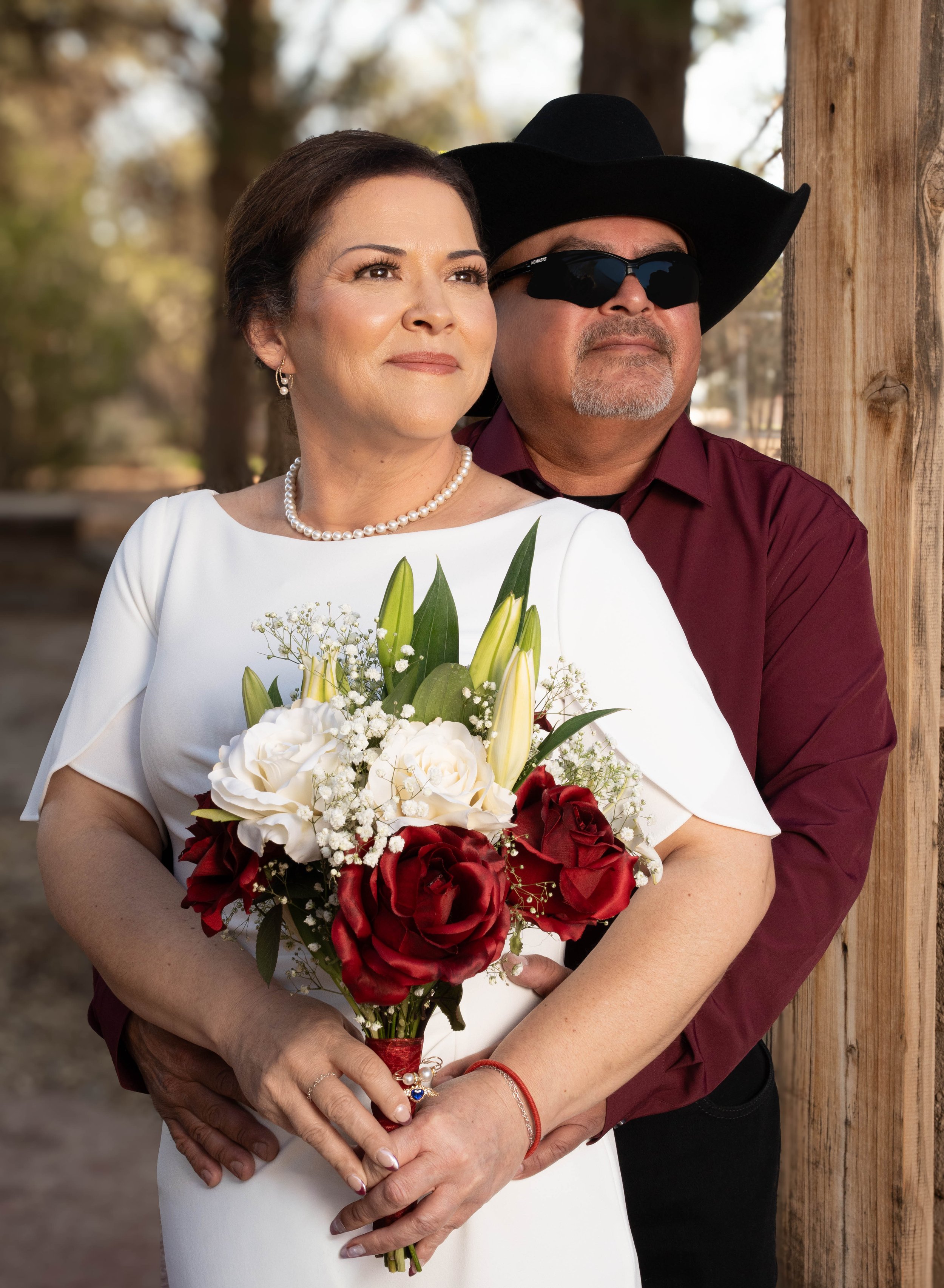 A woman in a white dress holding a bouquet of white and red roses, standing next to a man in sunglasses, a black hat, and a red shirt, outside near a wooden post.