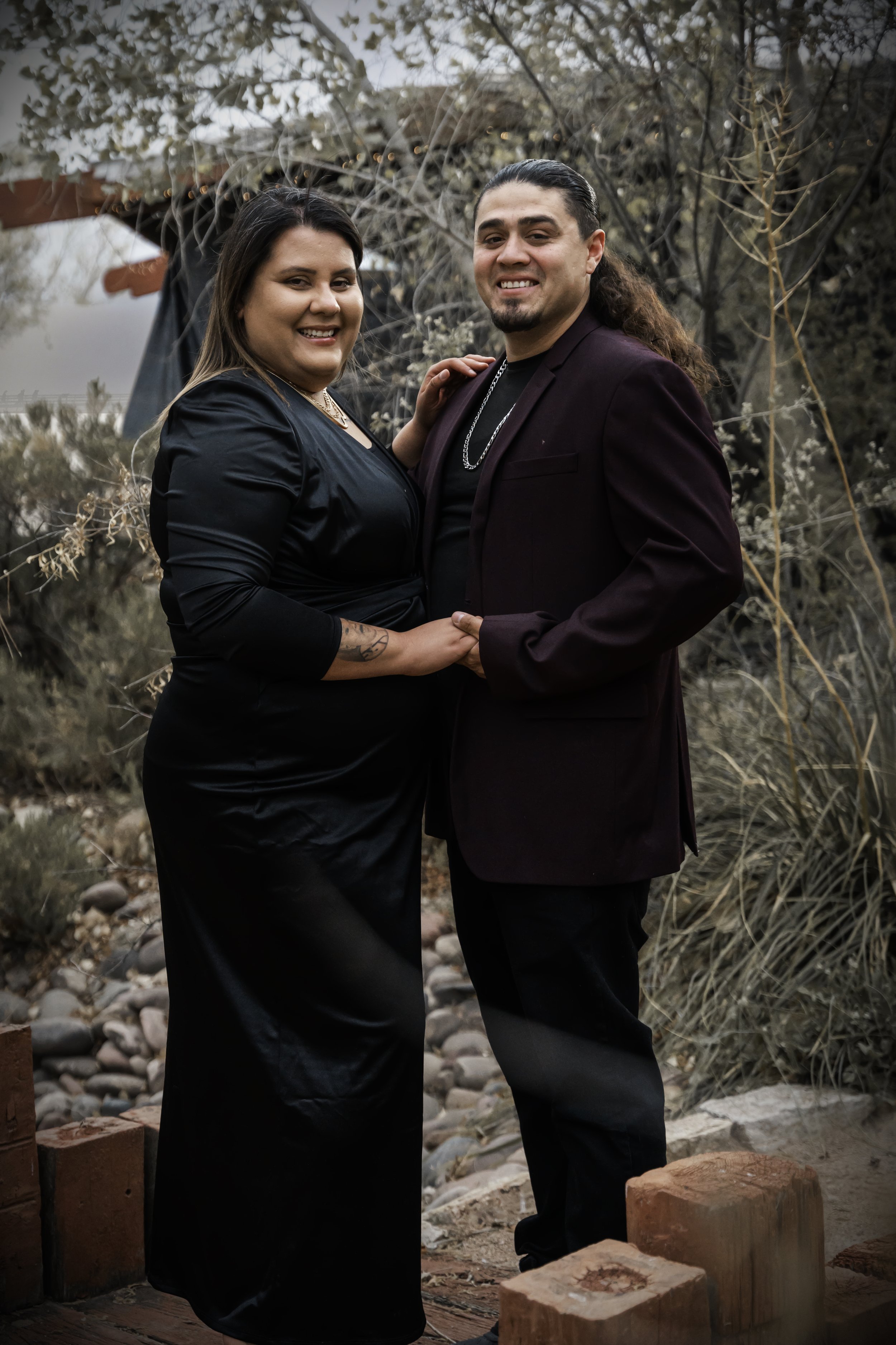 A couple dressed in formal attire holding hands and smiling outdoors in front of a wooden structure and trees.