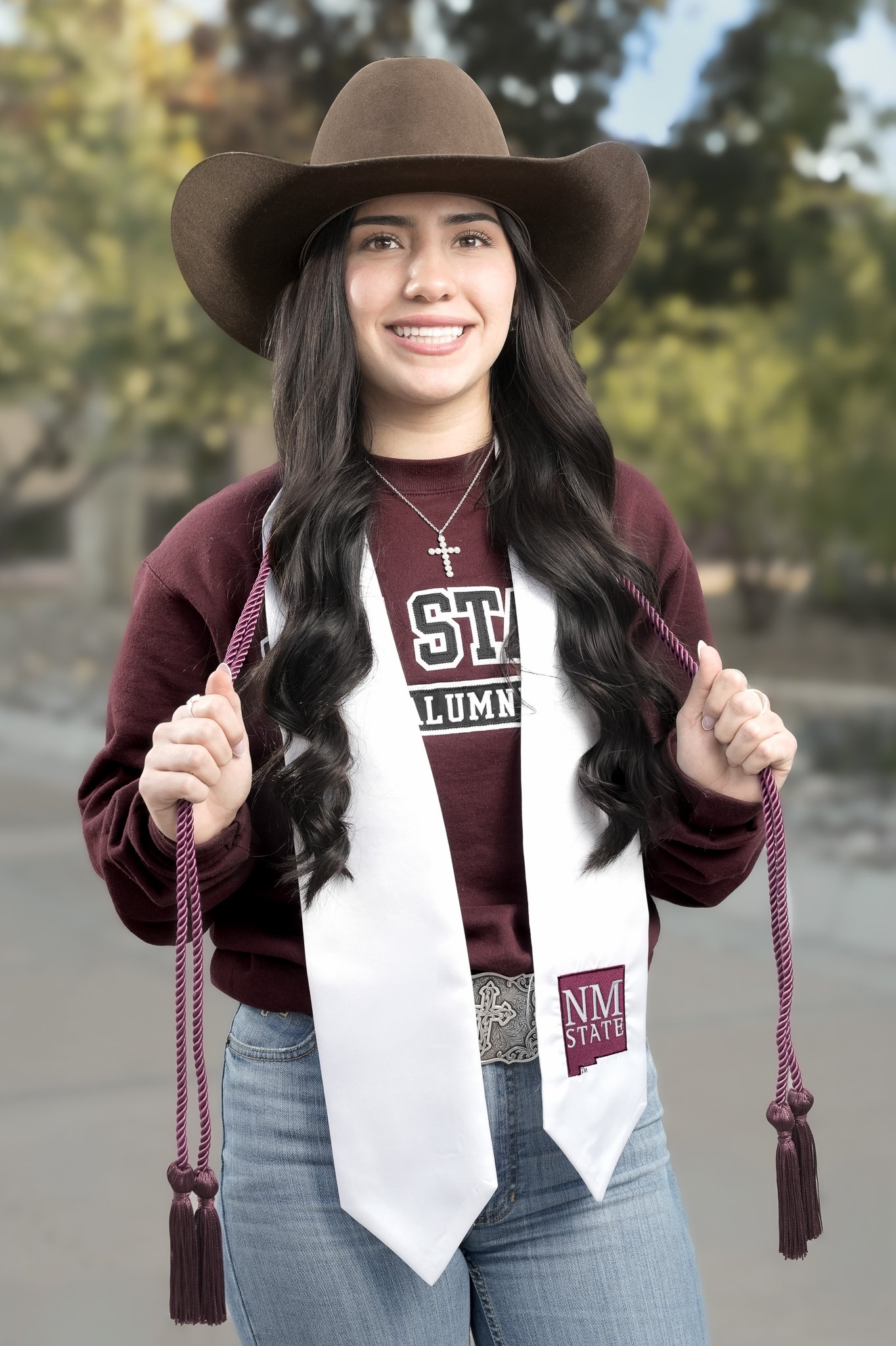Young woman in graduation cap and stole, wearing a maroon sweatshirt with 'STATE LUMNI' printed on it, holding the cords of her graduation cap, standing outdoors with trees in the background.
