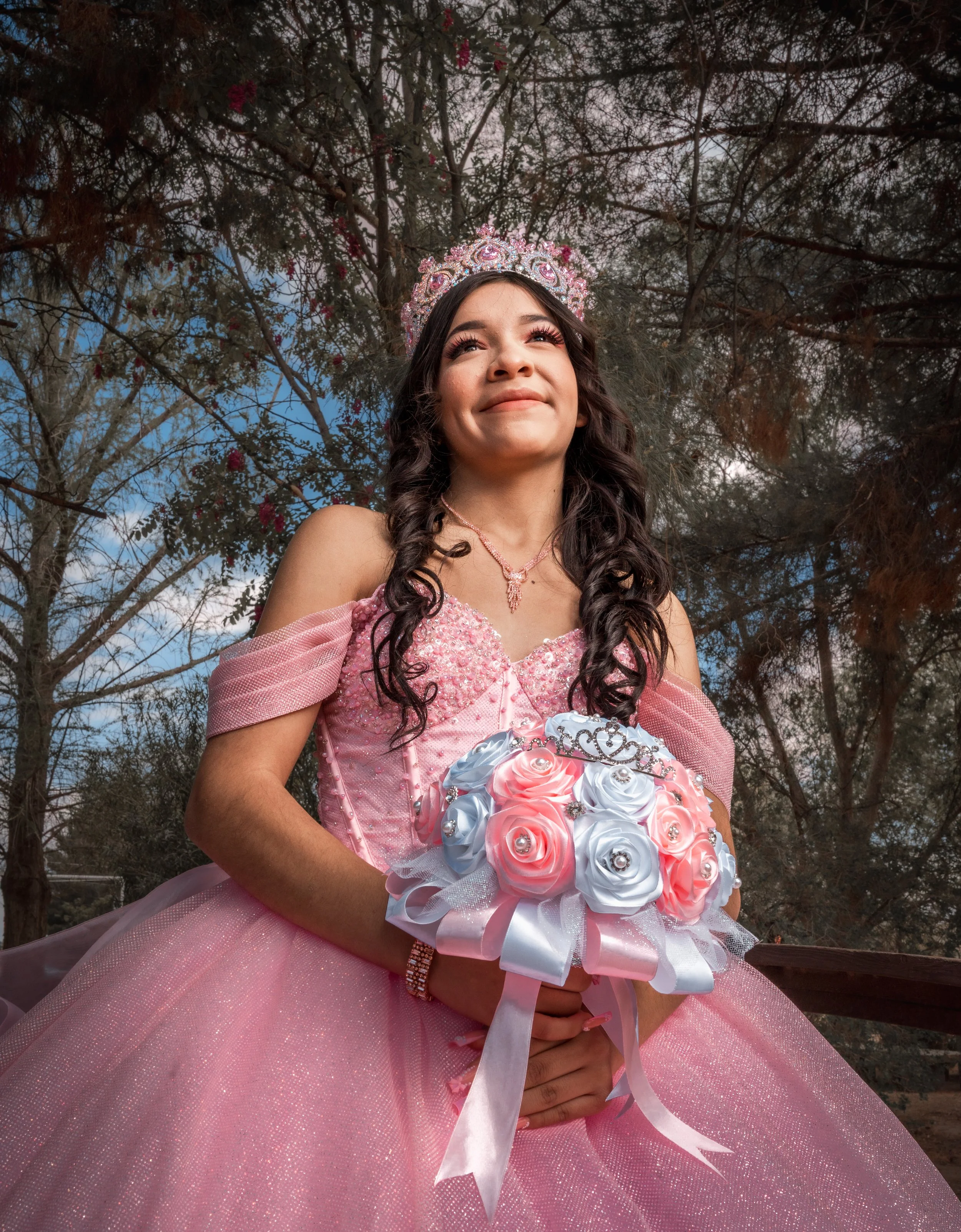 Young woman in a pink princess dress and tiara holding a bouquet of pink and white roses, standing outdoors with trees and a cloudy sky in the background.