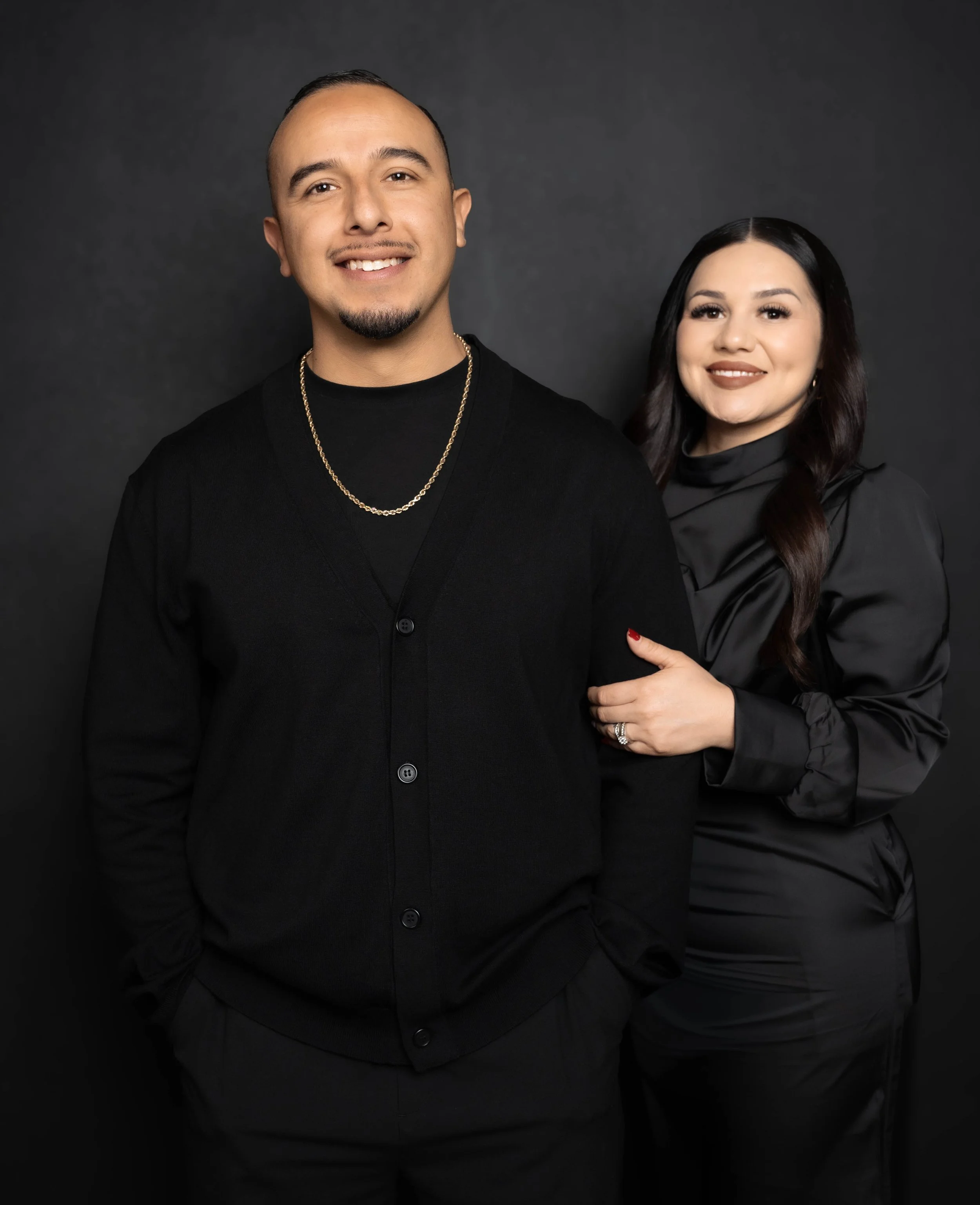 A man and woman posing together against a dark background, both dressed in black. The man is smiling, wearing a black cardigan, black shirt, gold chain, and has his hands in his pockets. The woman is smiling, with dark wavy hair, wearing a black satin blouse, with her arm around the man's arm and wearing a ring and red nail polish.