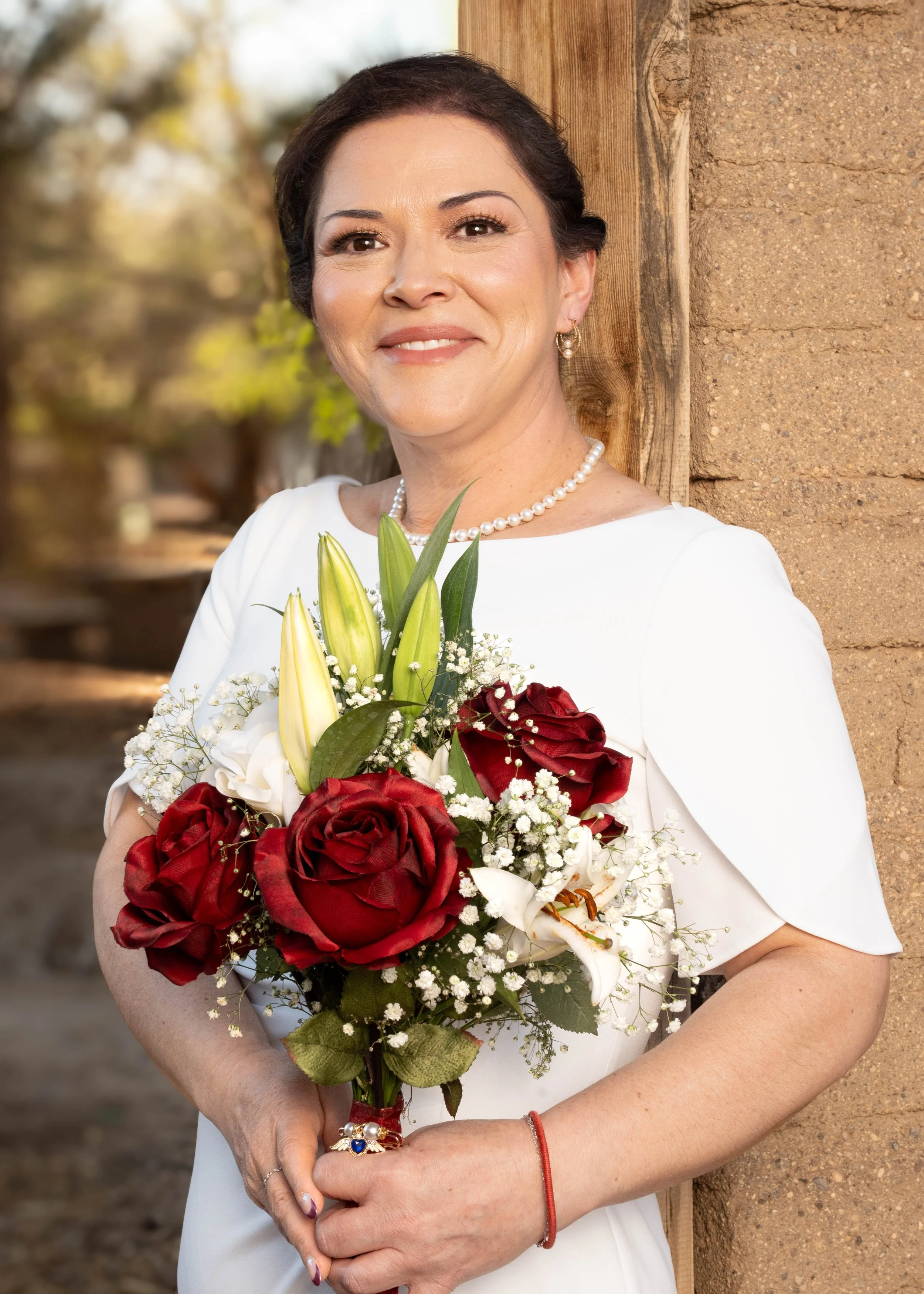 A woman in a white dress holding a bouquet of red and white flowers, standing outdoors near a wooden post with a blurred natural background.