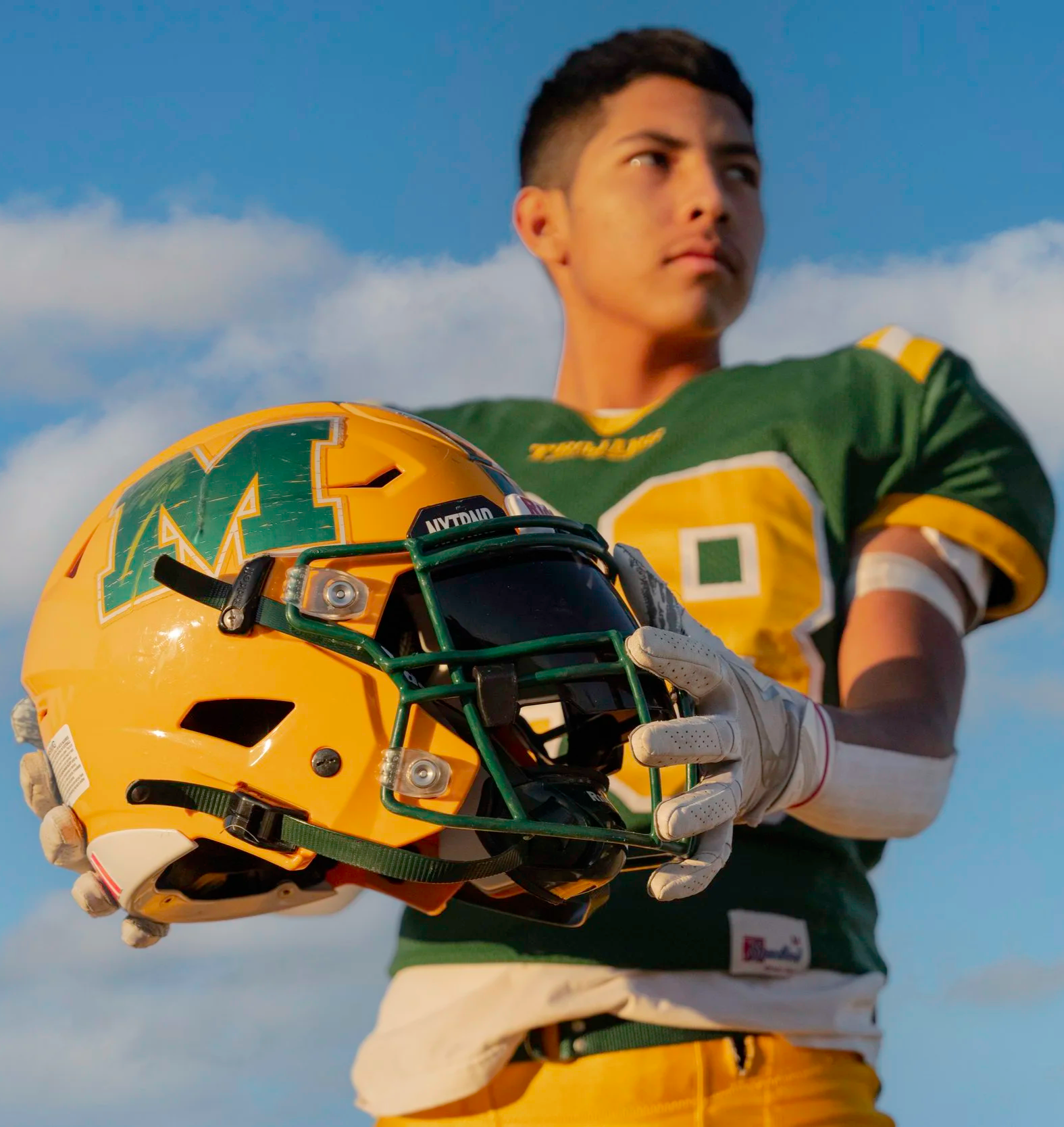 A young football player in a green jersey and yellow pants holds a yellow football helmet with a green logo and faceguard, outdoors with a blue sky and clouds in the background.