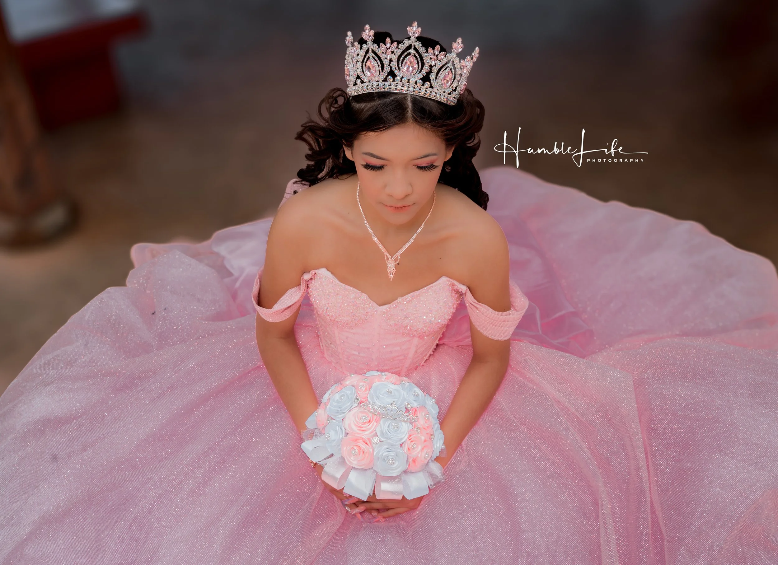 A young girl dressed as a princess in a pink ball gown, wearing a tiara, holding a bouquet of pastel pink and white roses, sitting with her head bowed.