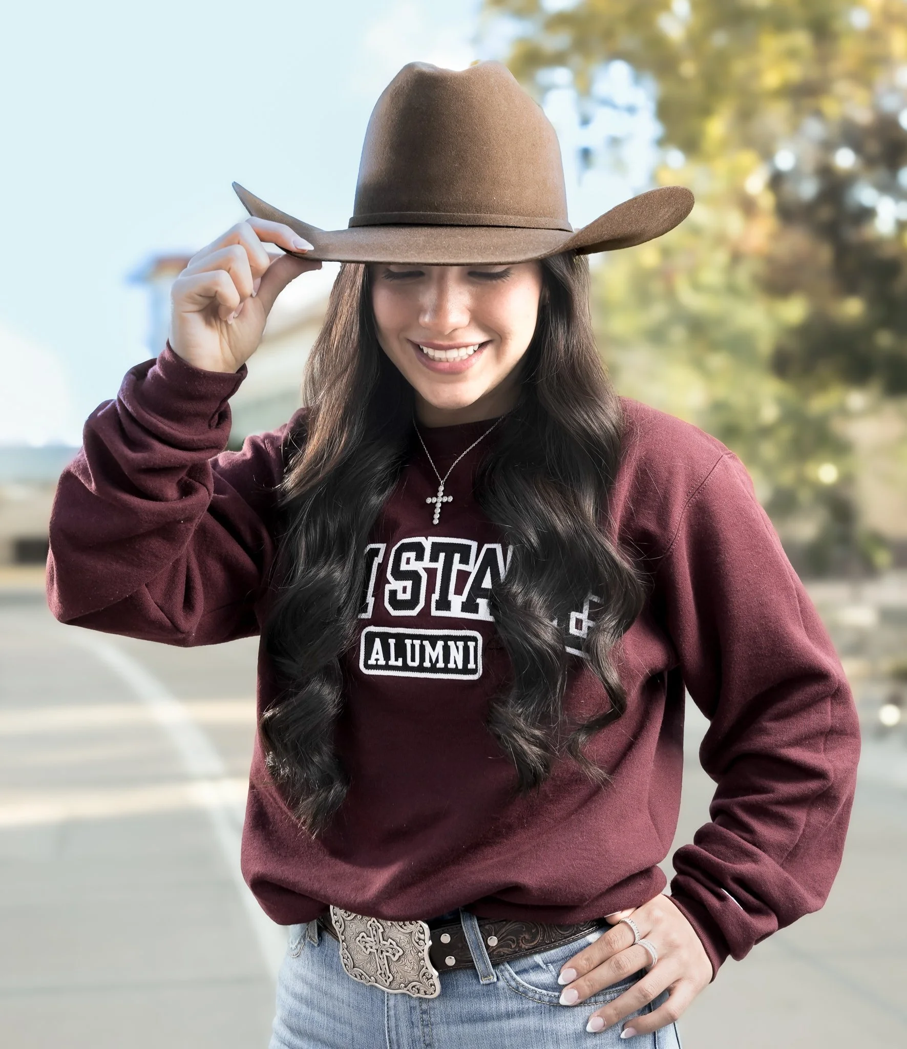 A smiling young woman wearing a cowboy hat, a maroon hoodie with 'ALUMNI' written on it, a cross necklace, and blue jeans, standing outdoors during daytime with trees in the background.