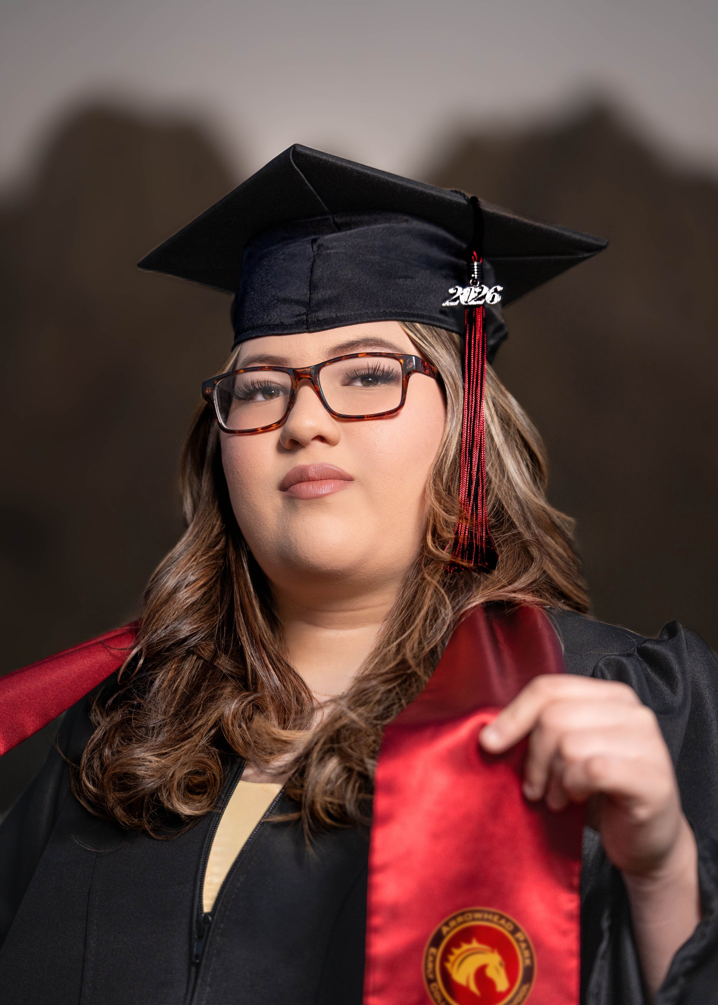 A woman wearing a black graduation cap and gown, holding a red stole with a yellow emblem, and wearing glasses.