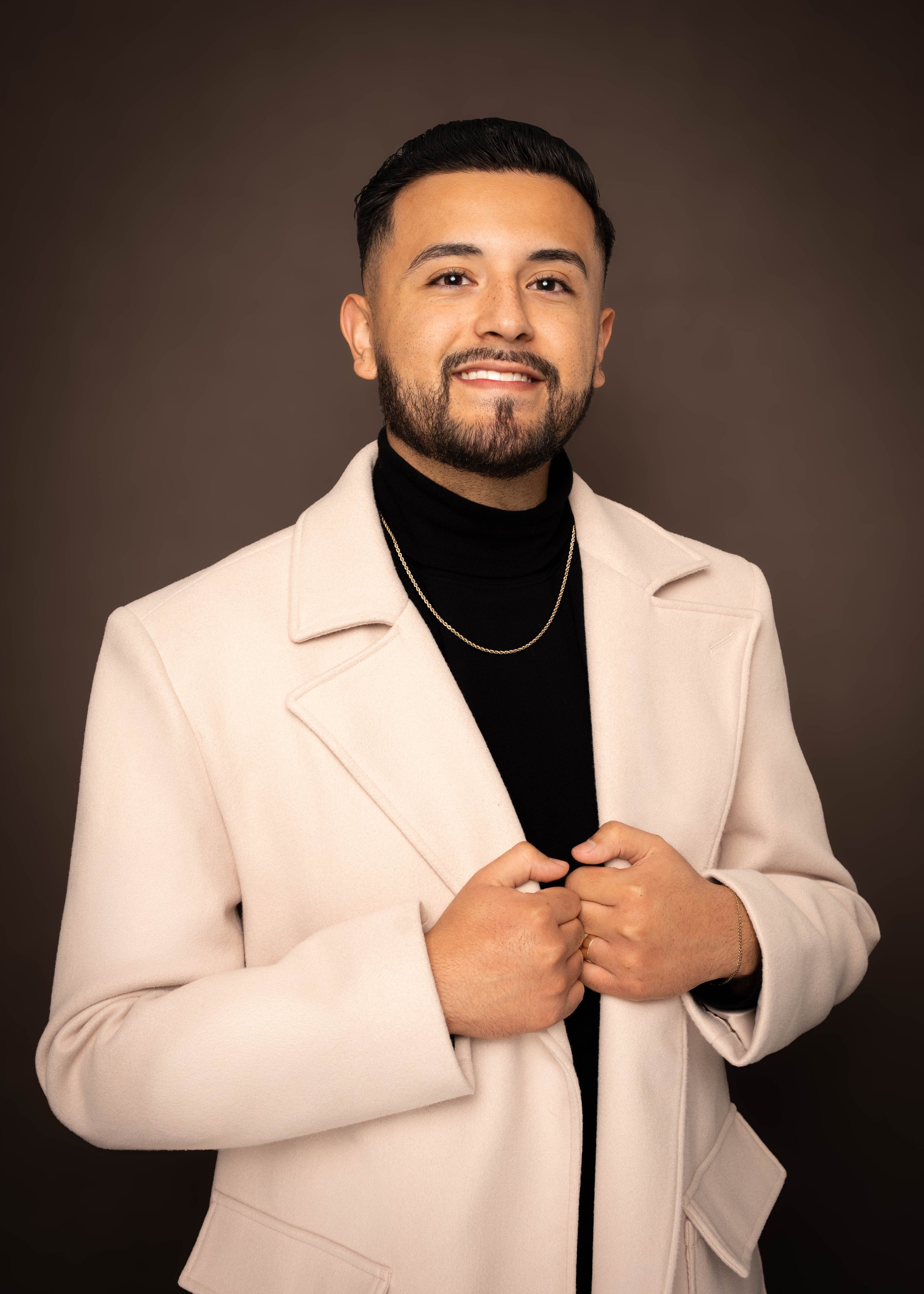 A man posing against a dark brown background, wearing a beige coat over a black turtleneck, with short dark hair and a beard, smiling and holding the lapels of his coat.