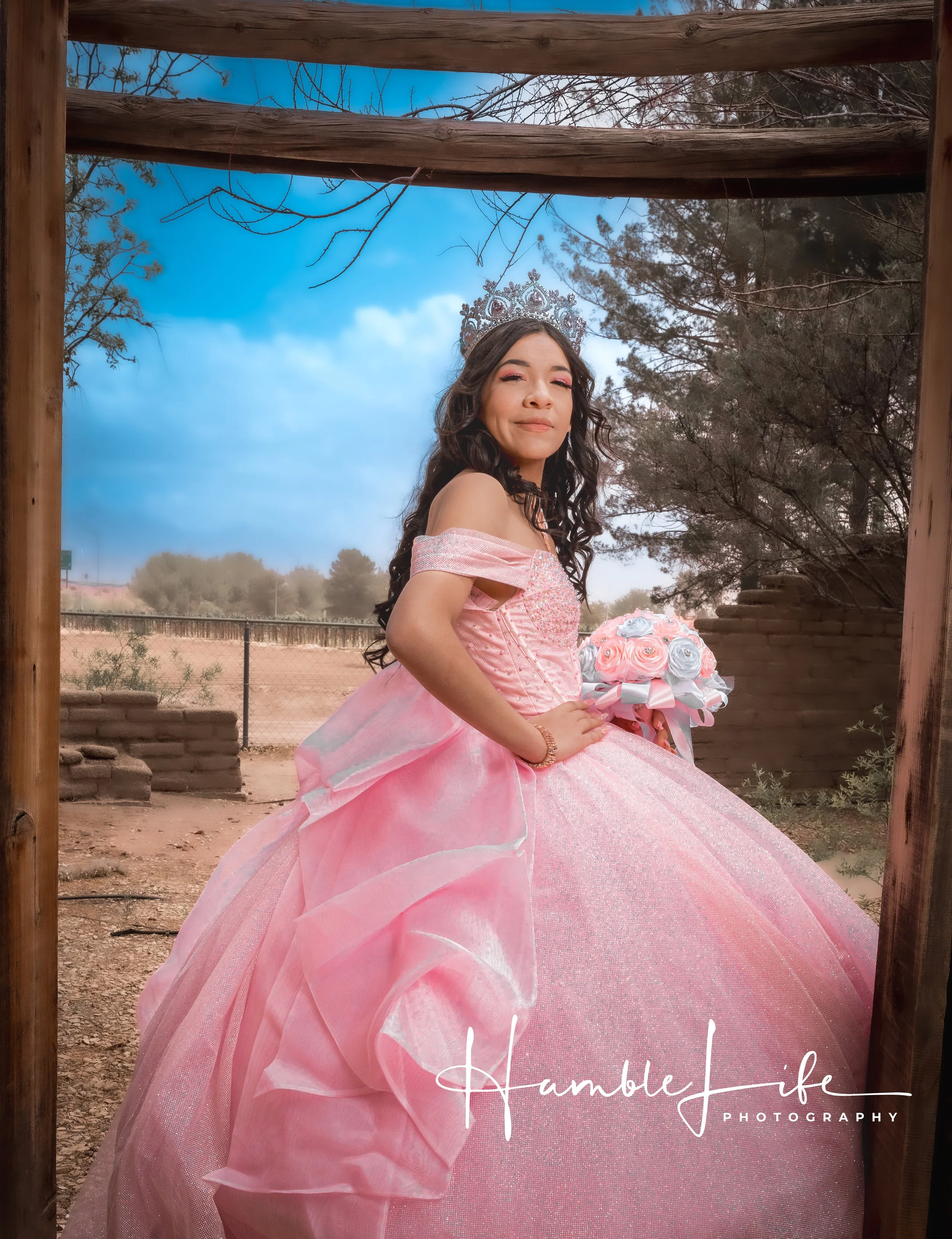 A young woman dressed in a pink princess gown with off-shoulder sleeves and a full skirt, standing outdoors under a wooden frame. She is wearing a tiara and holding a bouquet of pink and white roses. The background features trees, a fence, and a partly cloudy sky.