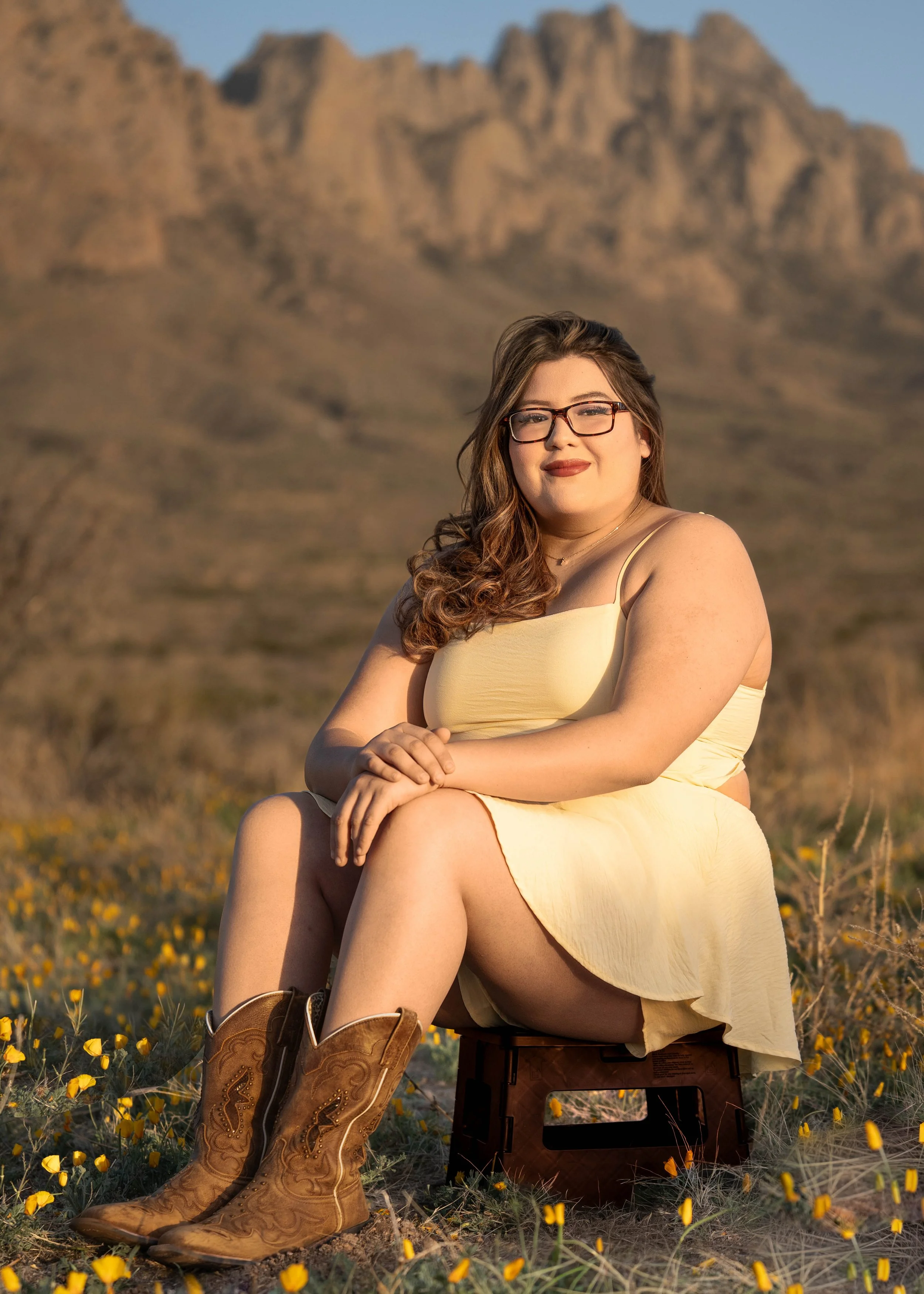 A woman wearing glasses and a yellow dress sitting on a small stool in a field of yellow flowers with mountains in the background.
