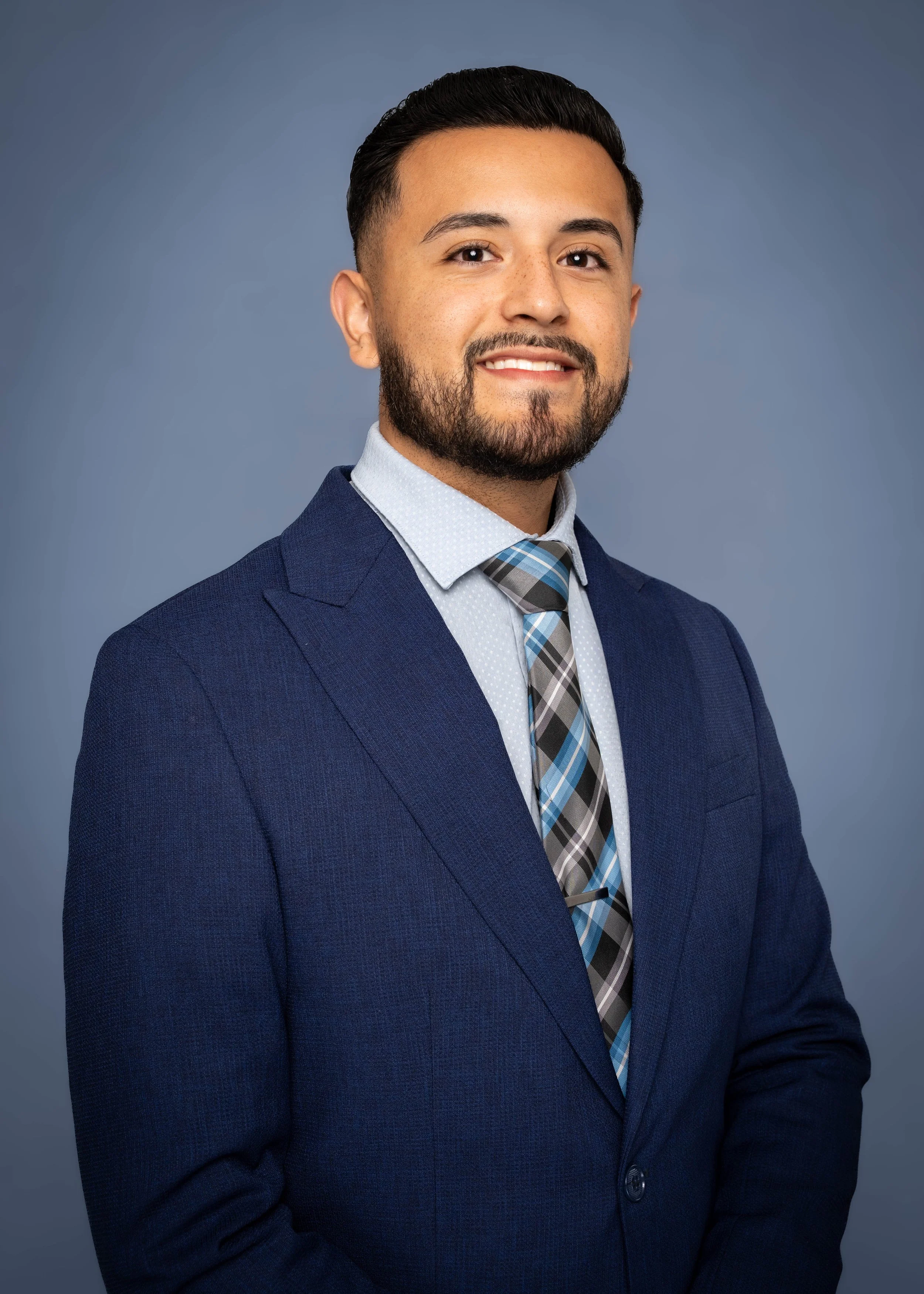 A professional man with dark hair and beard, wearing a navy blue suit, white shirt, and patterned tie, smiling against a plain gray background.