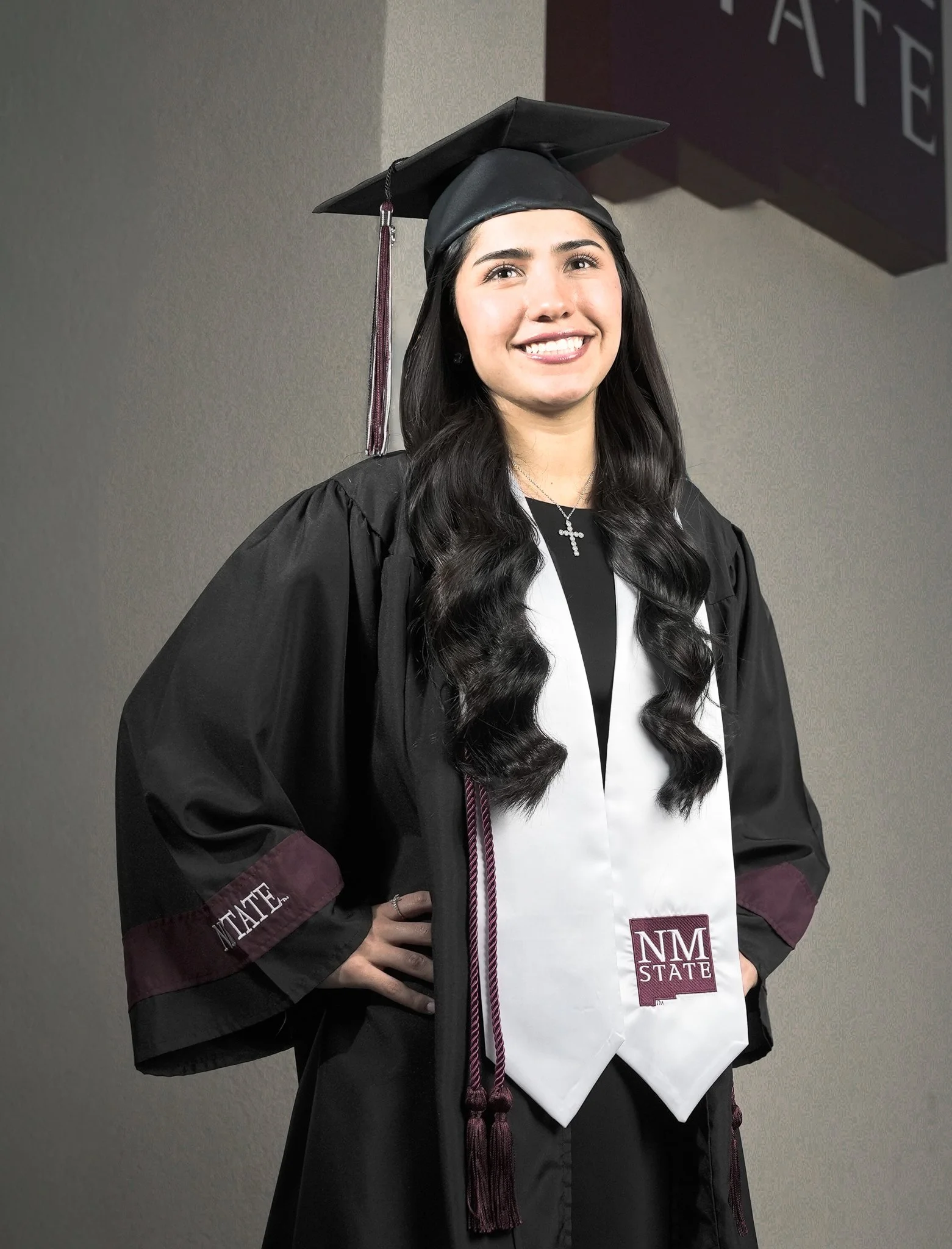 A young woman in a black graduation cap and gown, smiling, with long wavy black hair, standing indoors against a plain wall. She is wearing a white stole with the NM State logo and a necklace with a cross.