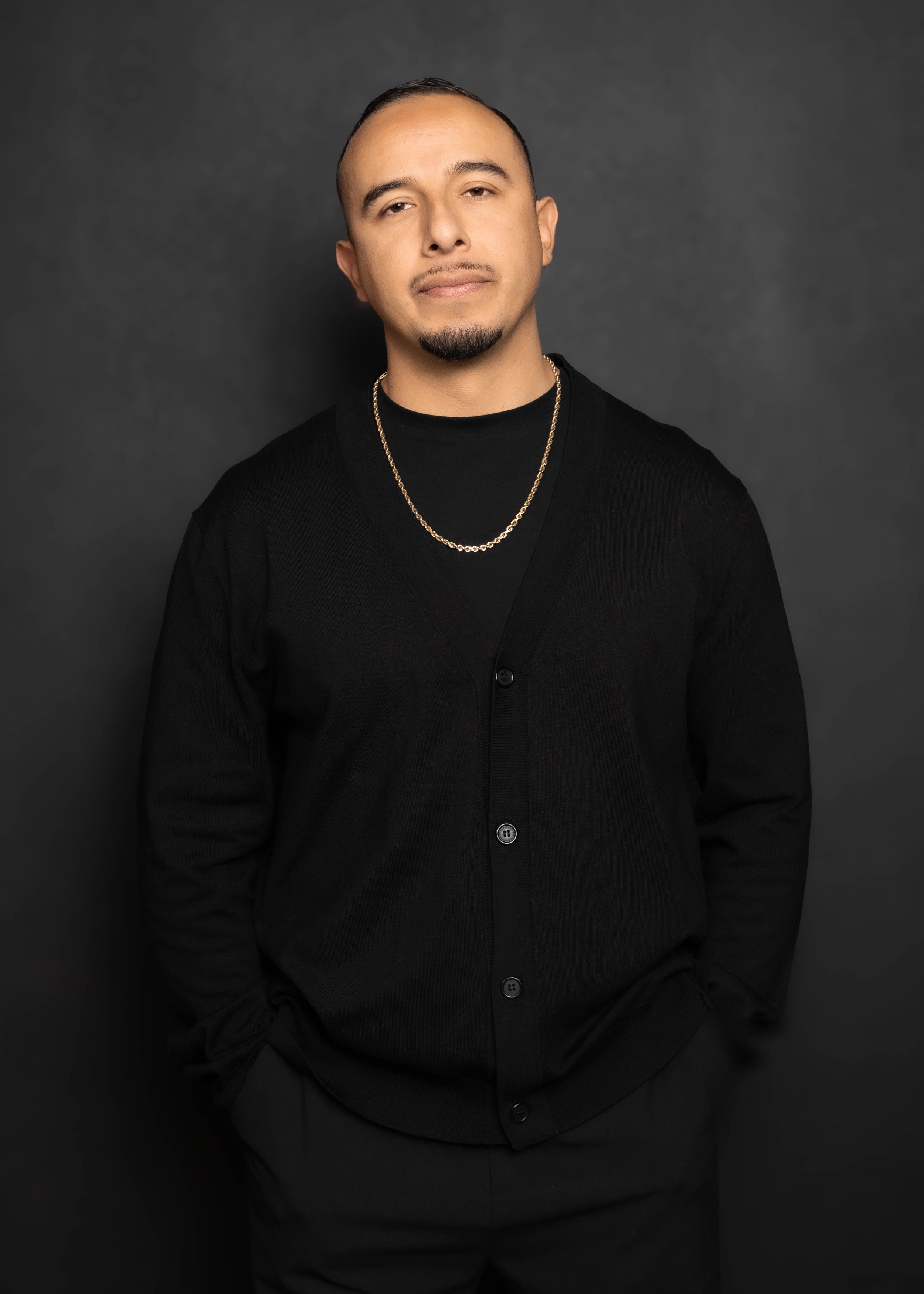 Portrait of a young man with short hair, light mustache and goatee, wearing a black cardigan over a black shirt, gold chain necklace, and standing against a dark gray background.
