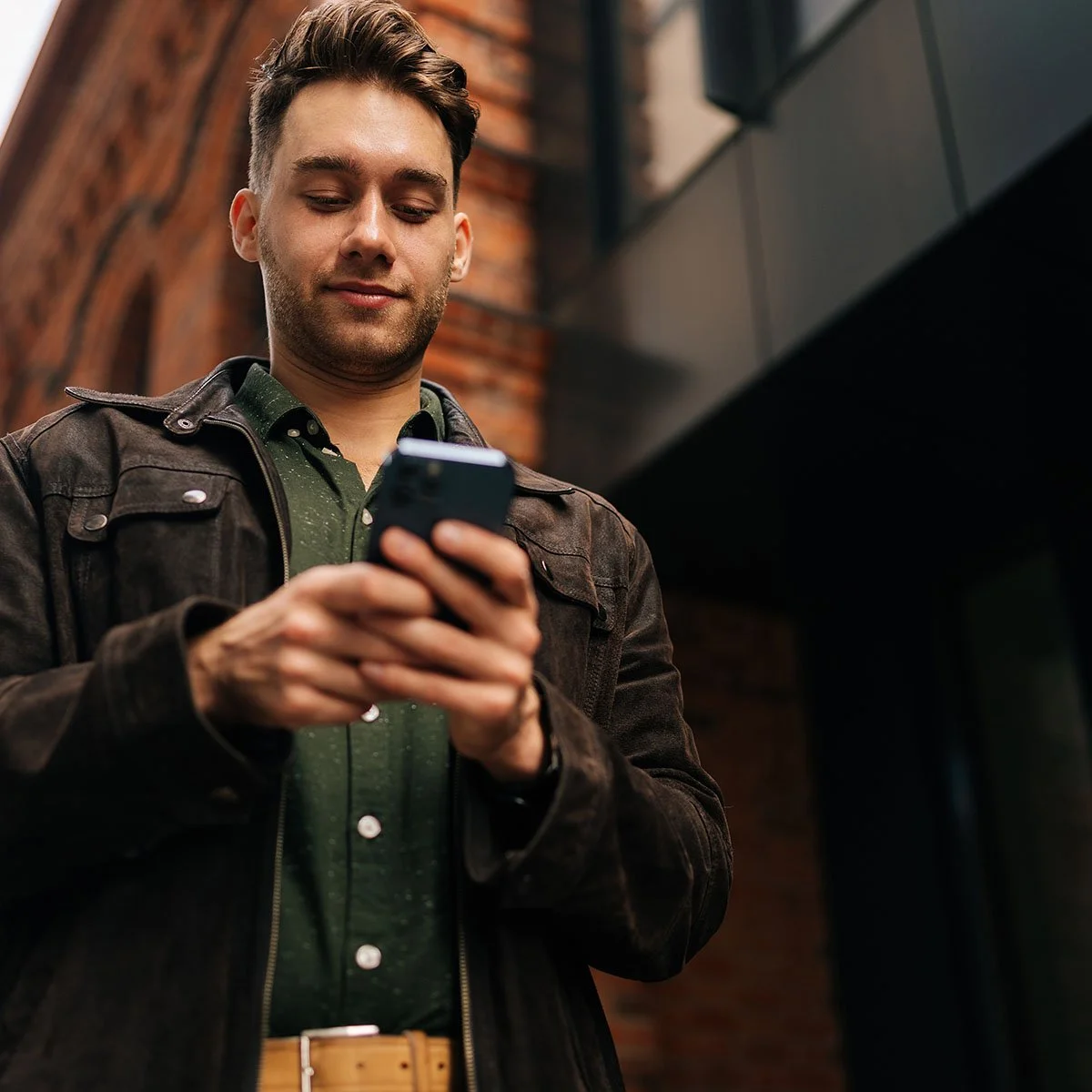 A young man with short brown hair looking at his smartphone, standing outdoors near a brick wall and modern black architectural elements.