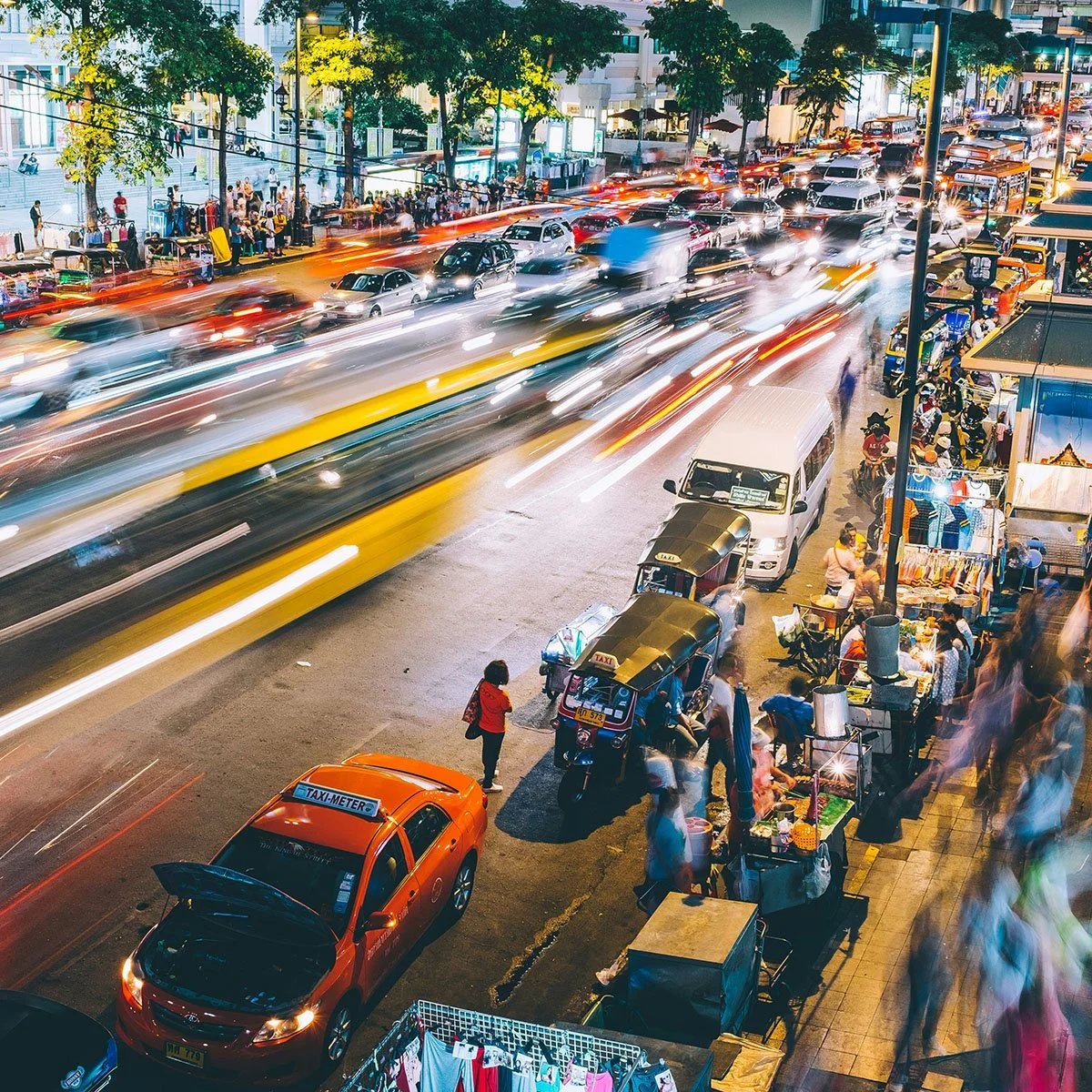 Nighttime busy city street with moving vehicles creating light trails, parked cars and tuk-tuks, street vendors and shoppers on the sidewalk, and high-rise buildings and trees in the background.