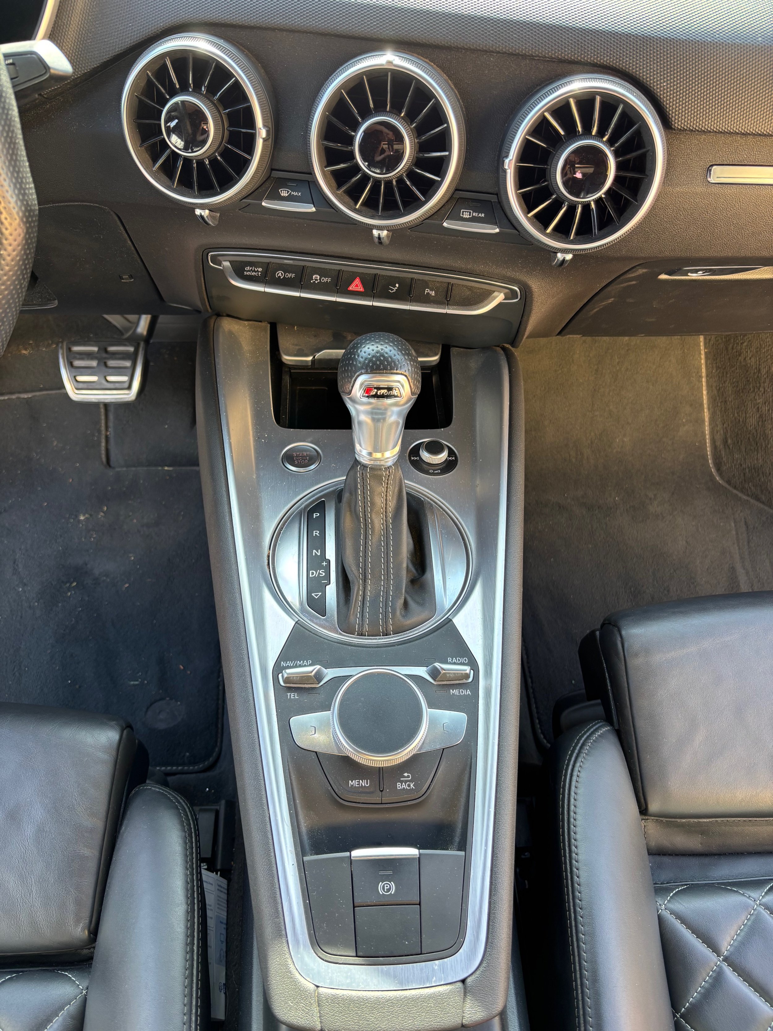 Tableau de bord d'une voiture avec volant, console centrale avec levier de vitesse, commandes pour navigation, radio, menu, et boutons pour la gestion du véhicule.