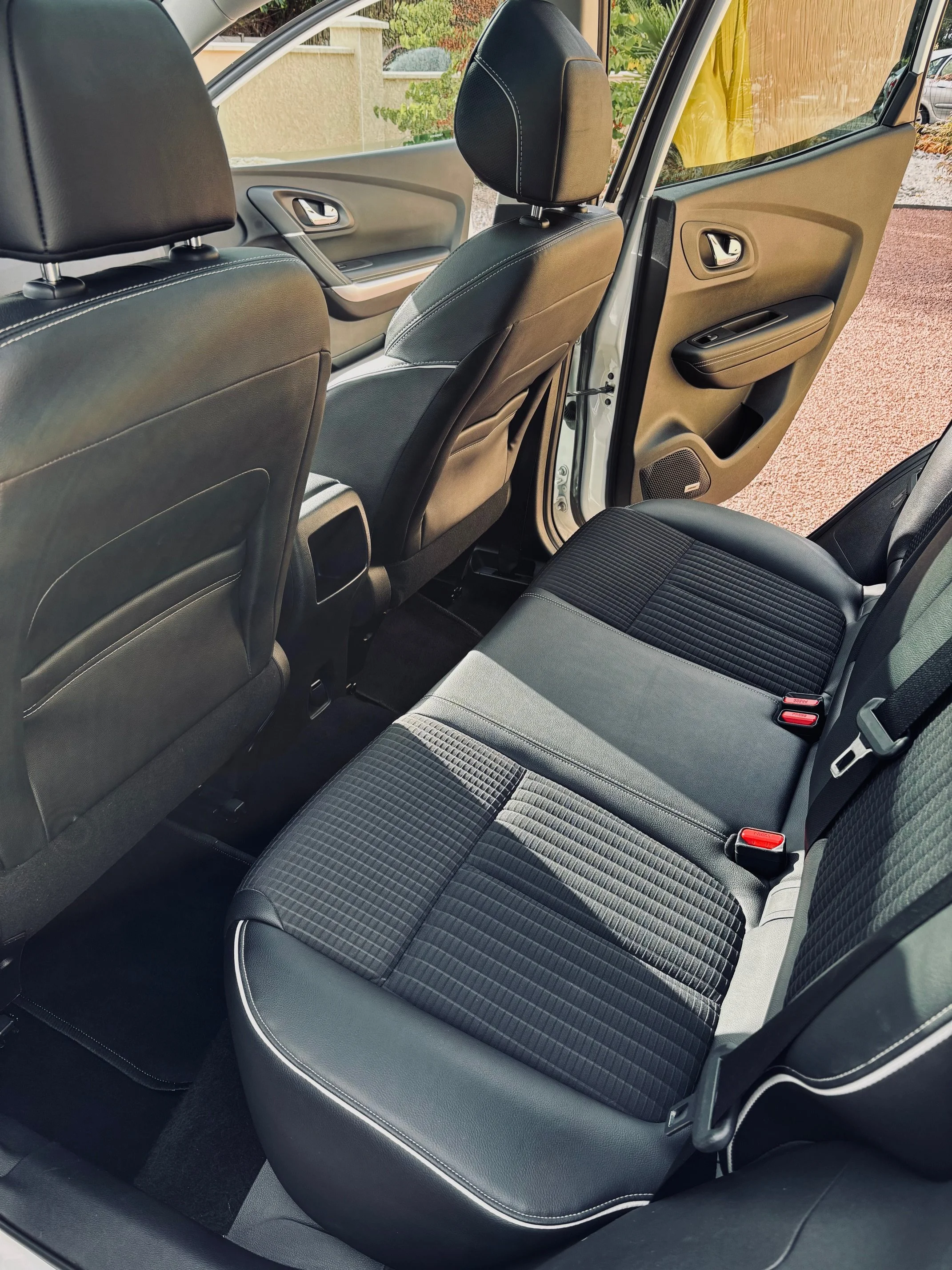 Intérieur d'une voiture, vue des sièges arrière, avec des sièges en tissu noir à motifs, porte arrière ouverte, environnement extérieur visible par la fenêtre.